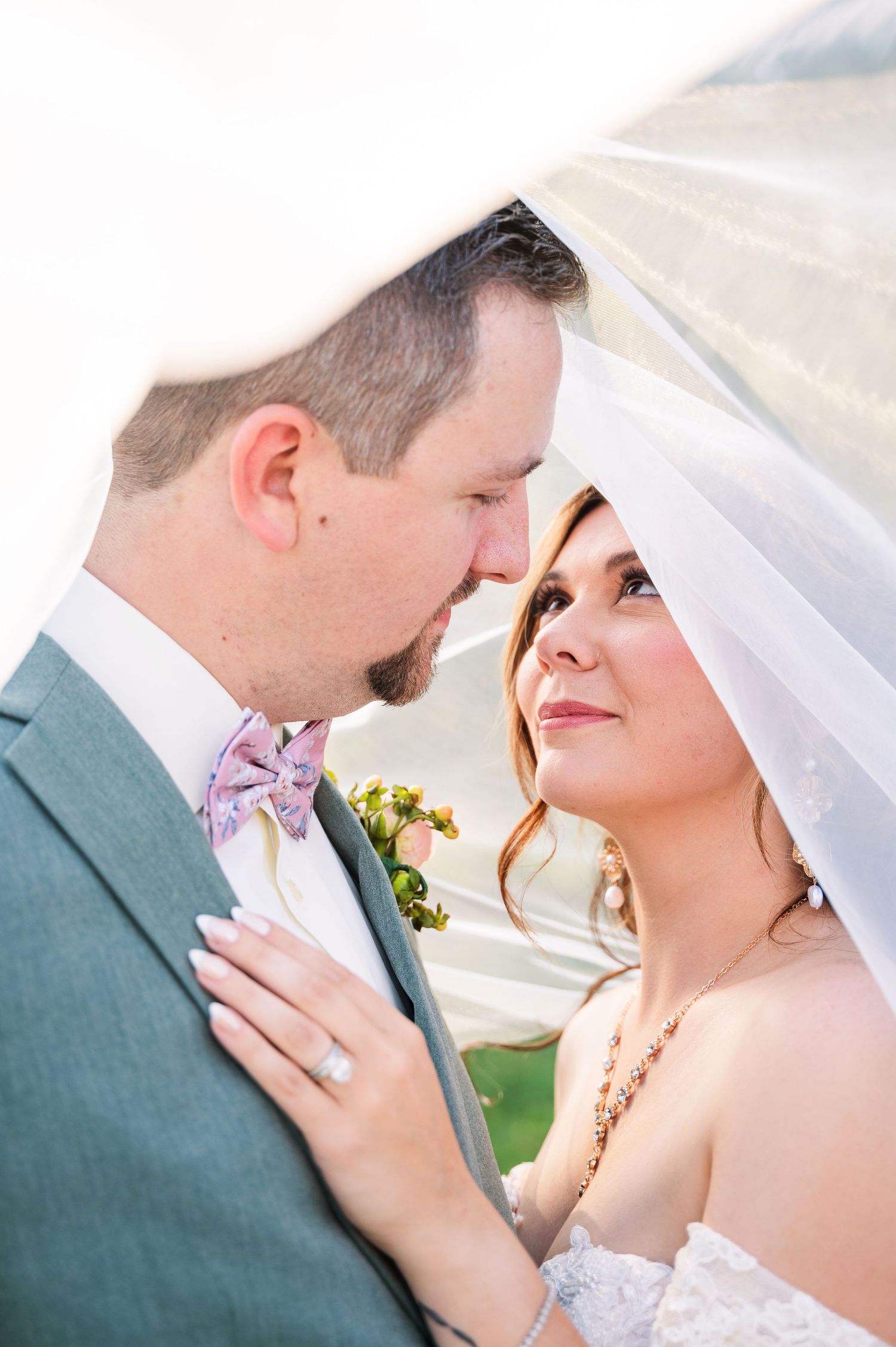 Bride and groom share an intimate moment under a veil, groom in gray suit with floral bow tie, bride in lace dress.