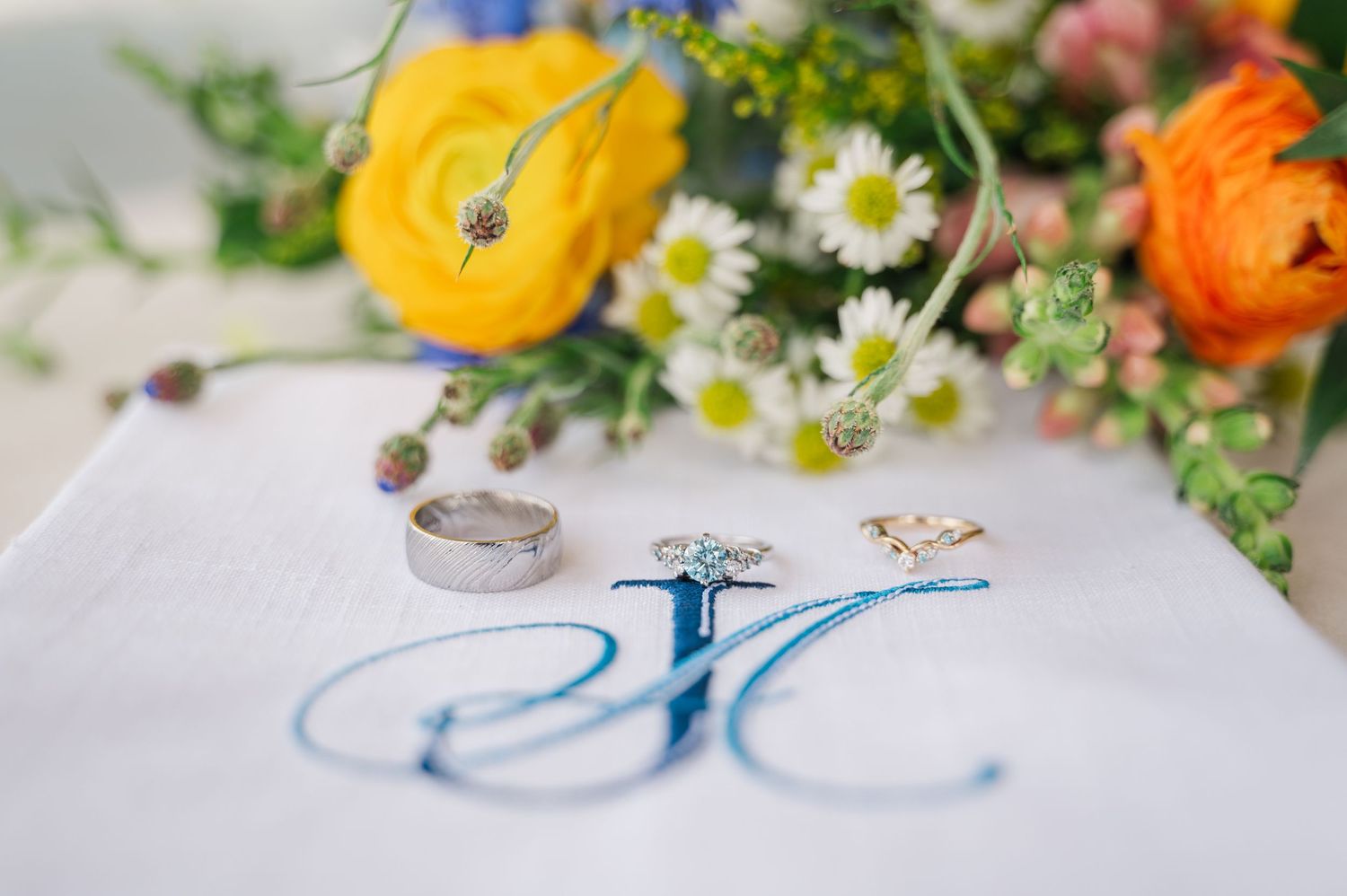 Wedding rings and earrings on embroidered monogram handkerchief with colorful floral bouquet featuring wildflower bouquet.