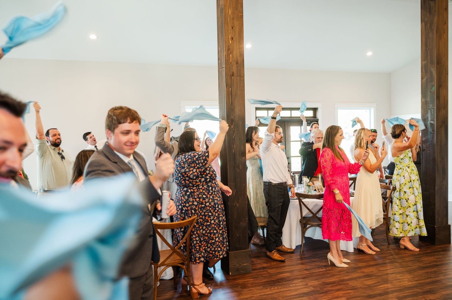 Wedding guests wave blue napkins on the dance floor inside a rustic wooden reception venue.