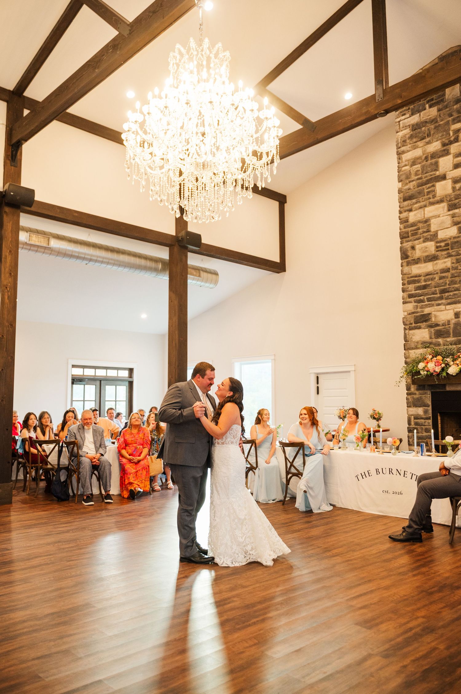 Bride and groom share a first dance beneath a crystal chandelier in a rustic wedding venue with wood beams and guests watching.