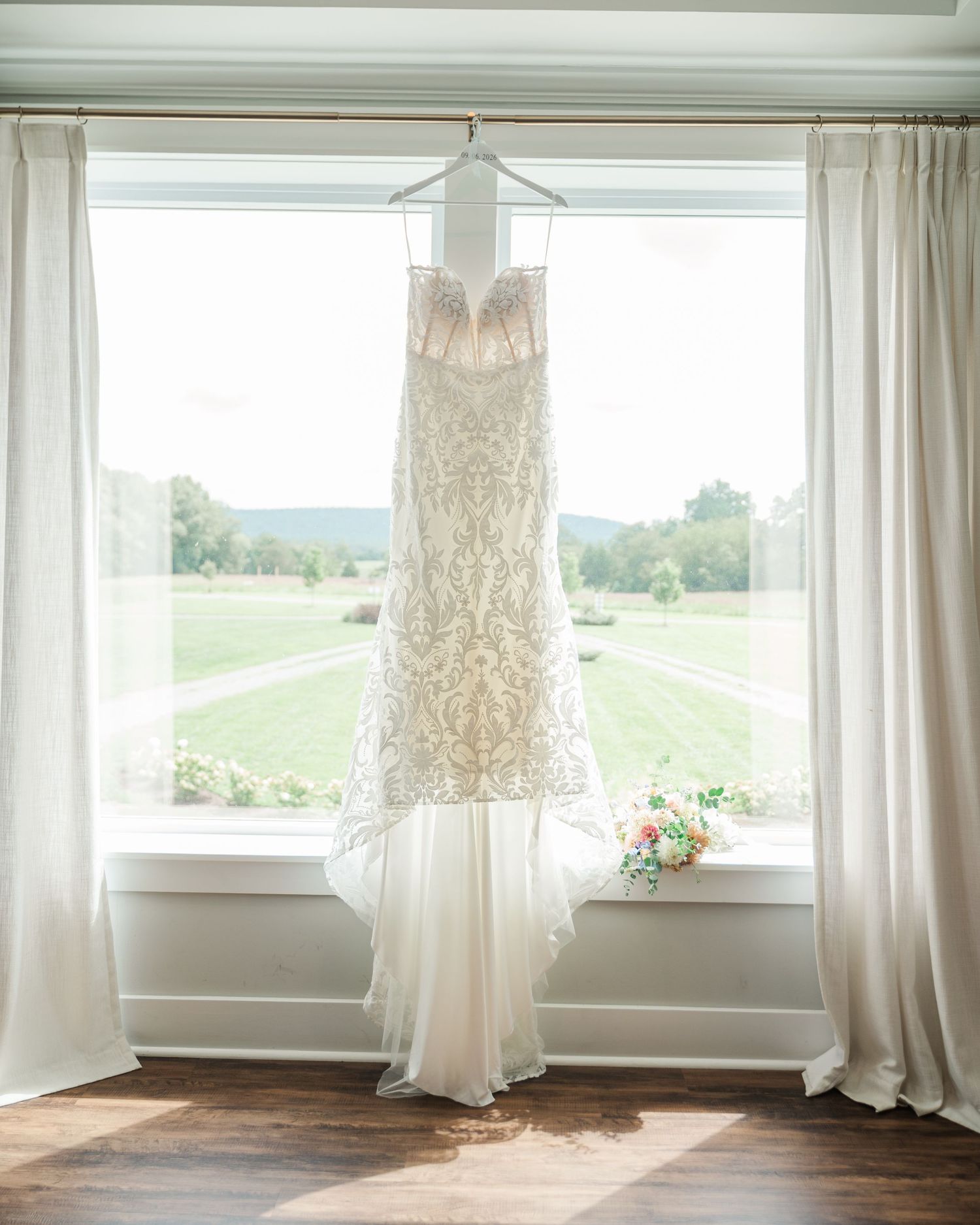 Elegant lace wedding dress hanging in a sunlit window with sheer curtains and a colorful bridal bouquet on the sill.