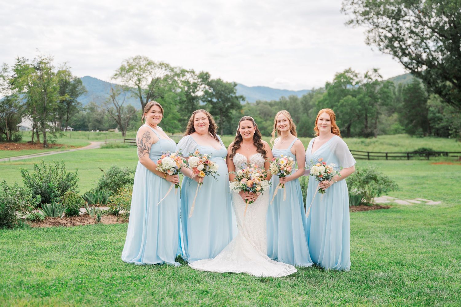 Bride in white gown with four bridesmaids in light blue dresses holding bouquets on green lawn with mountain backdrop.