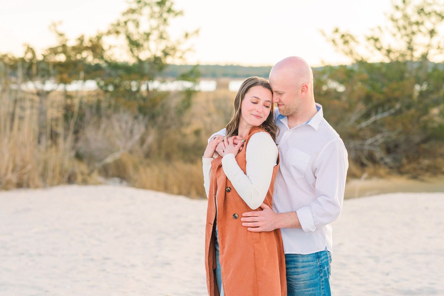 Couple embracing on sandy beach at golden hour, woman in rust vest and white top, man in light blue shirt and jeans.