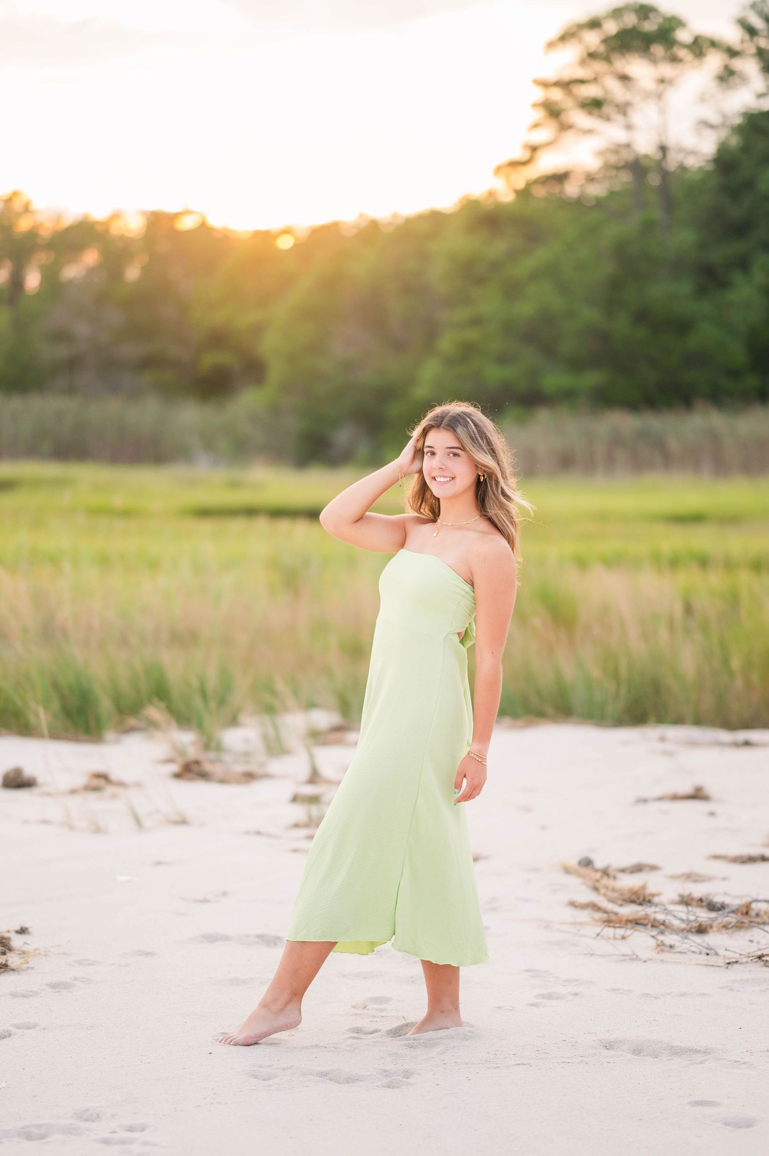 Young woman in mint green strapless dress walking barefoot on sandy beach at golden sunset.