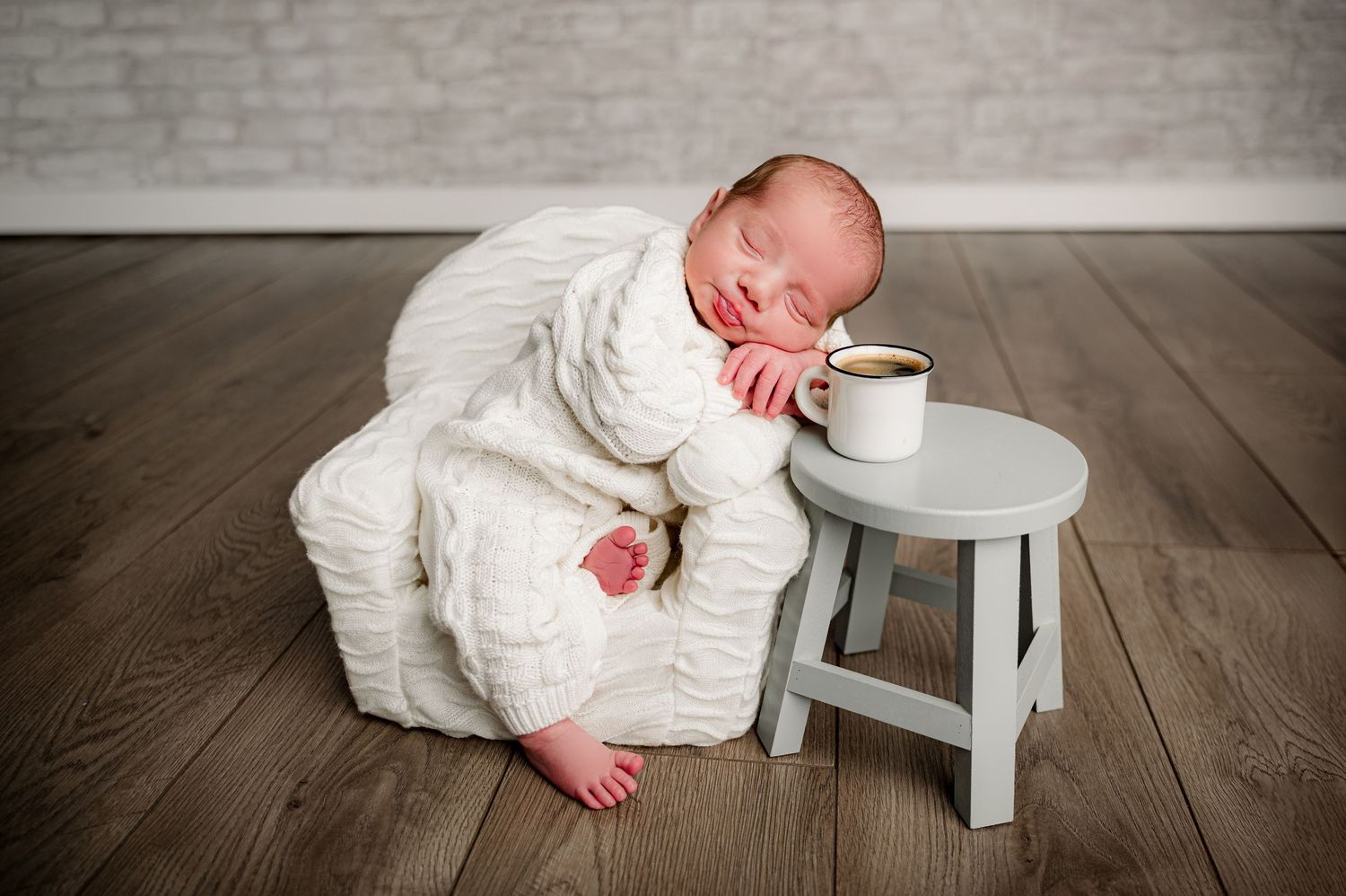 Newborn baby in white knit outfit posed next to a tiny gray stool with a coffee cup for studio newborn session.