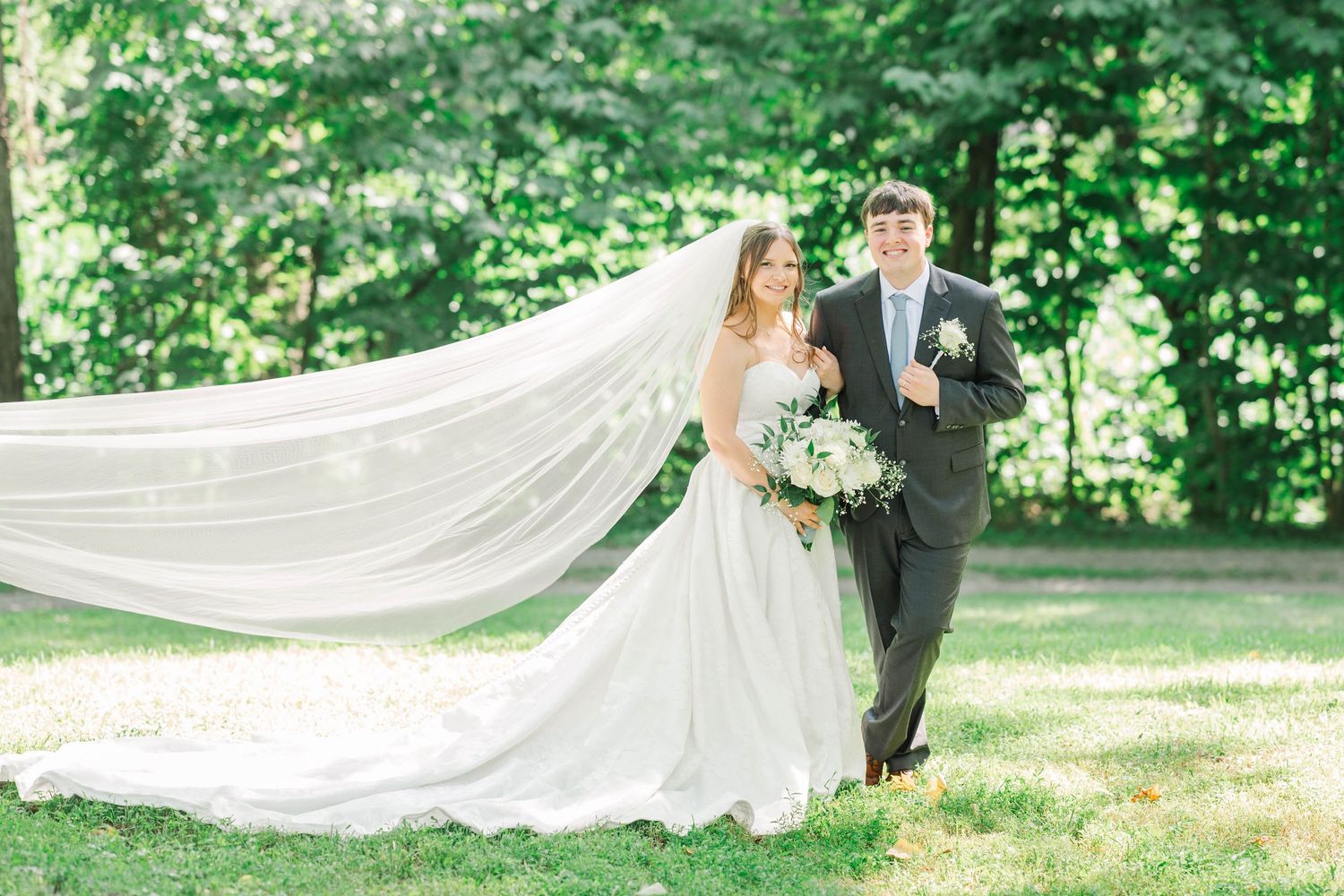 Wedding couple poses in sunlit garden with flowing white veil.