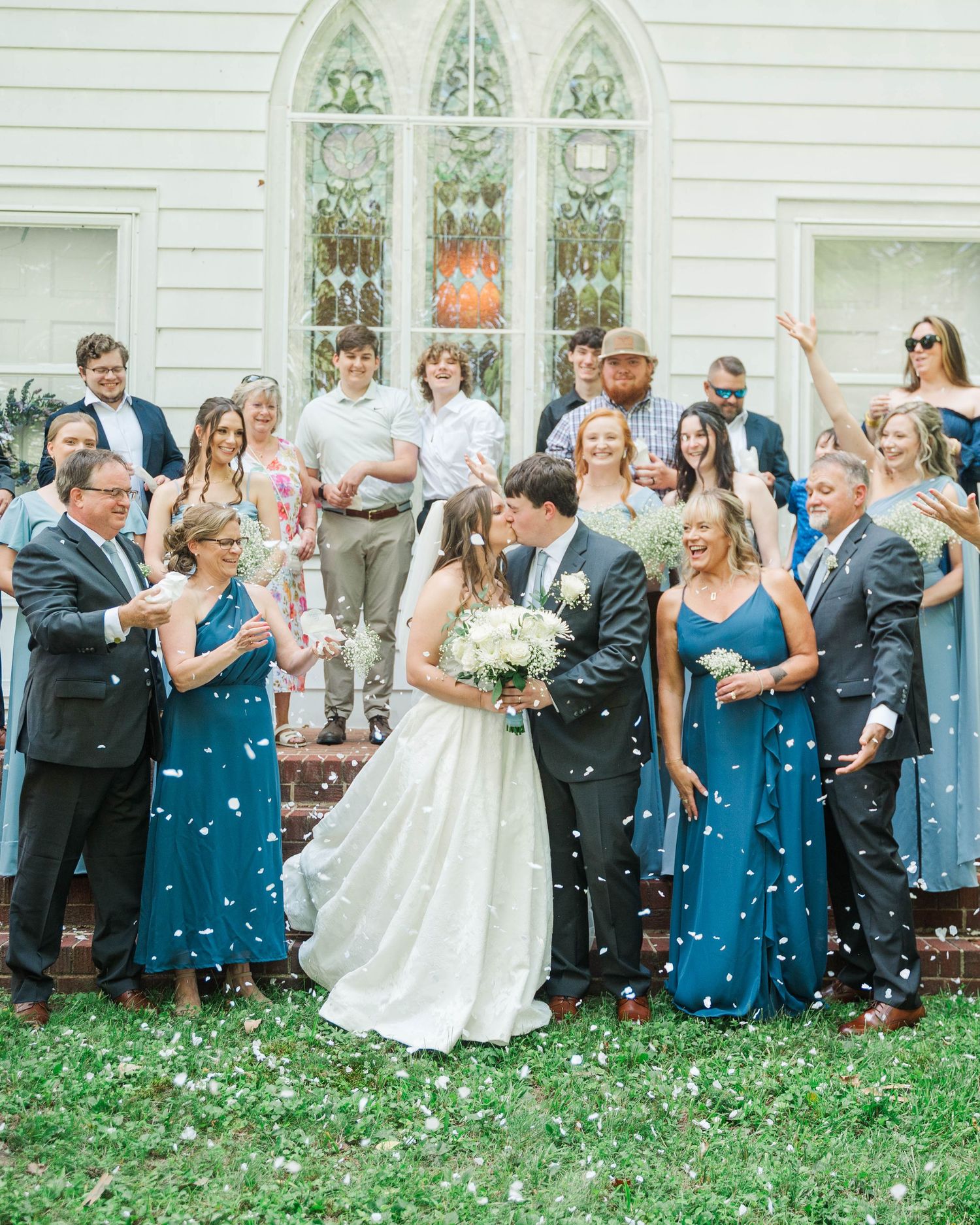 Wedding party and guests celebrate outside a white church as confetti falls.