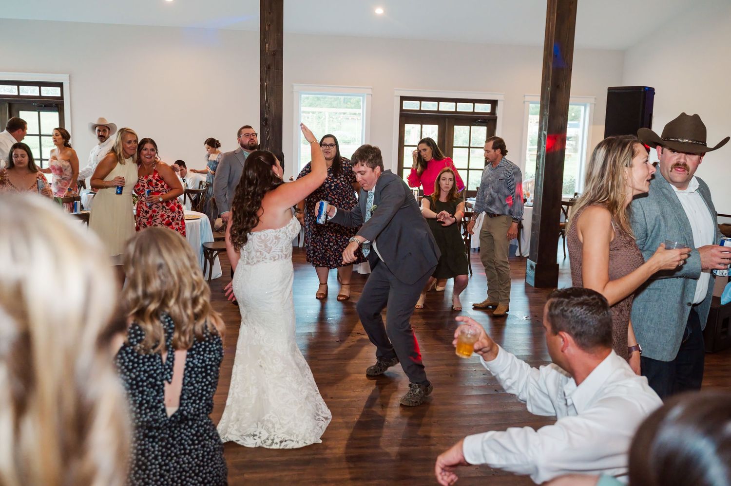 Bride in a white lace gown and groom in a gray suit dance as wedding guests clap and cheer in a rustic hall with wood floors.