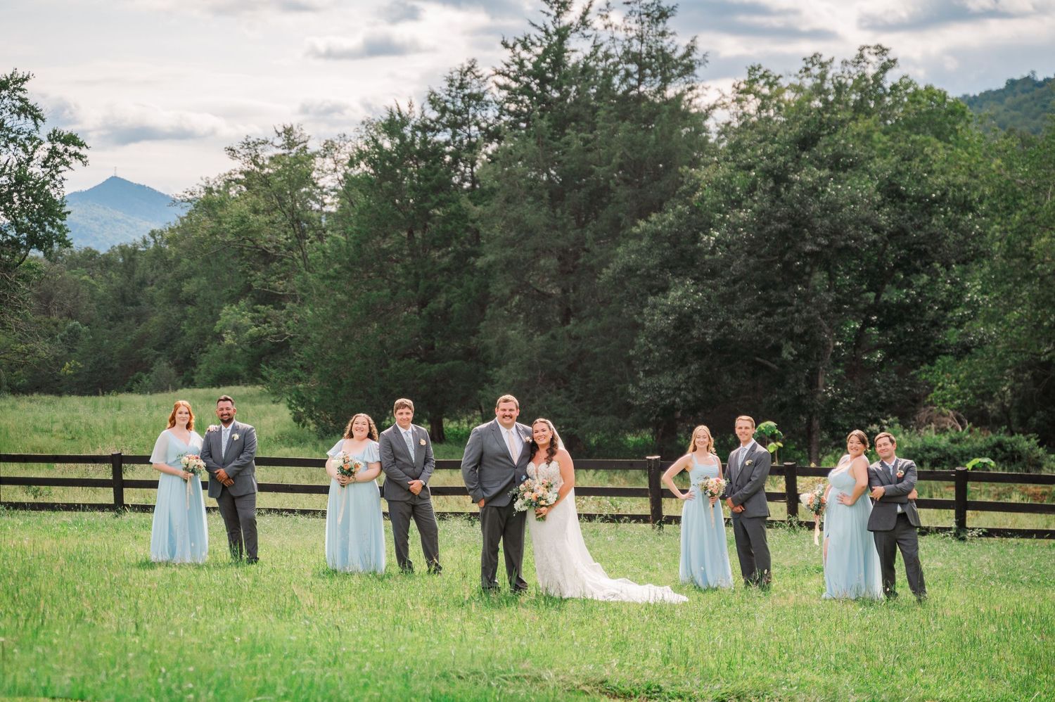 Wedding party poses in a lush green field with mountains and fence backdrop, bridesmaids in blue gowns.