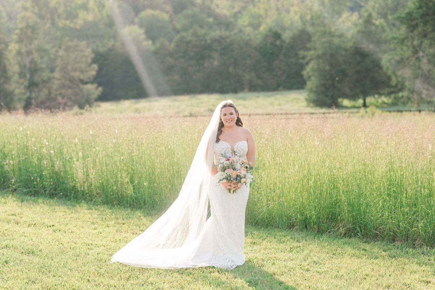 Bride in white gown and veil standing in a sunlit meadow holding a pastel bouquet on her wedding day.