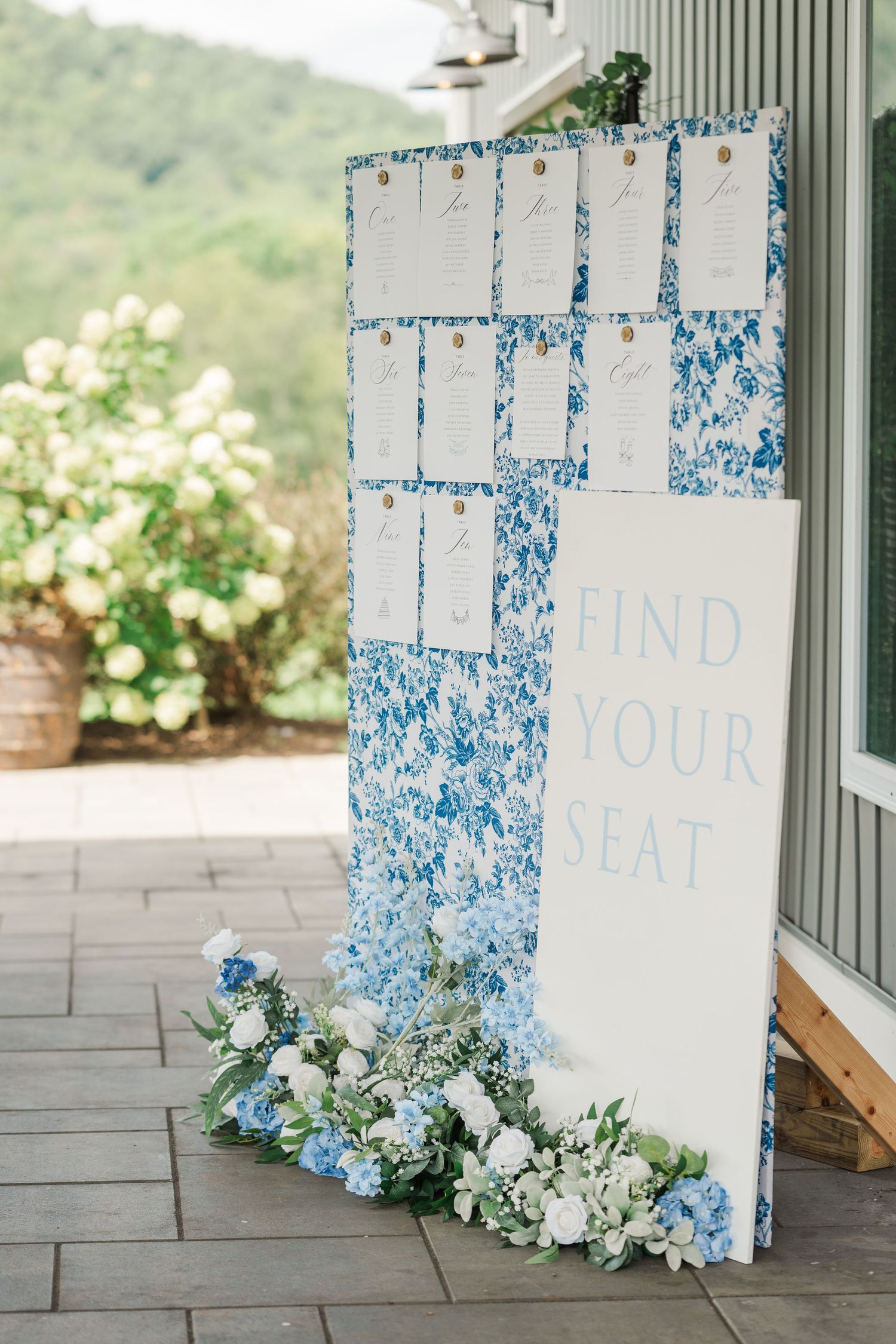 Blue floral wedding seating chart display with white find your seat sign adorned with hydrangeas and greenery at base.