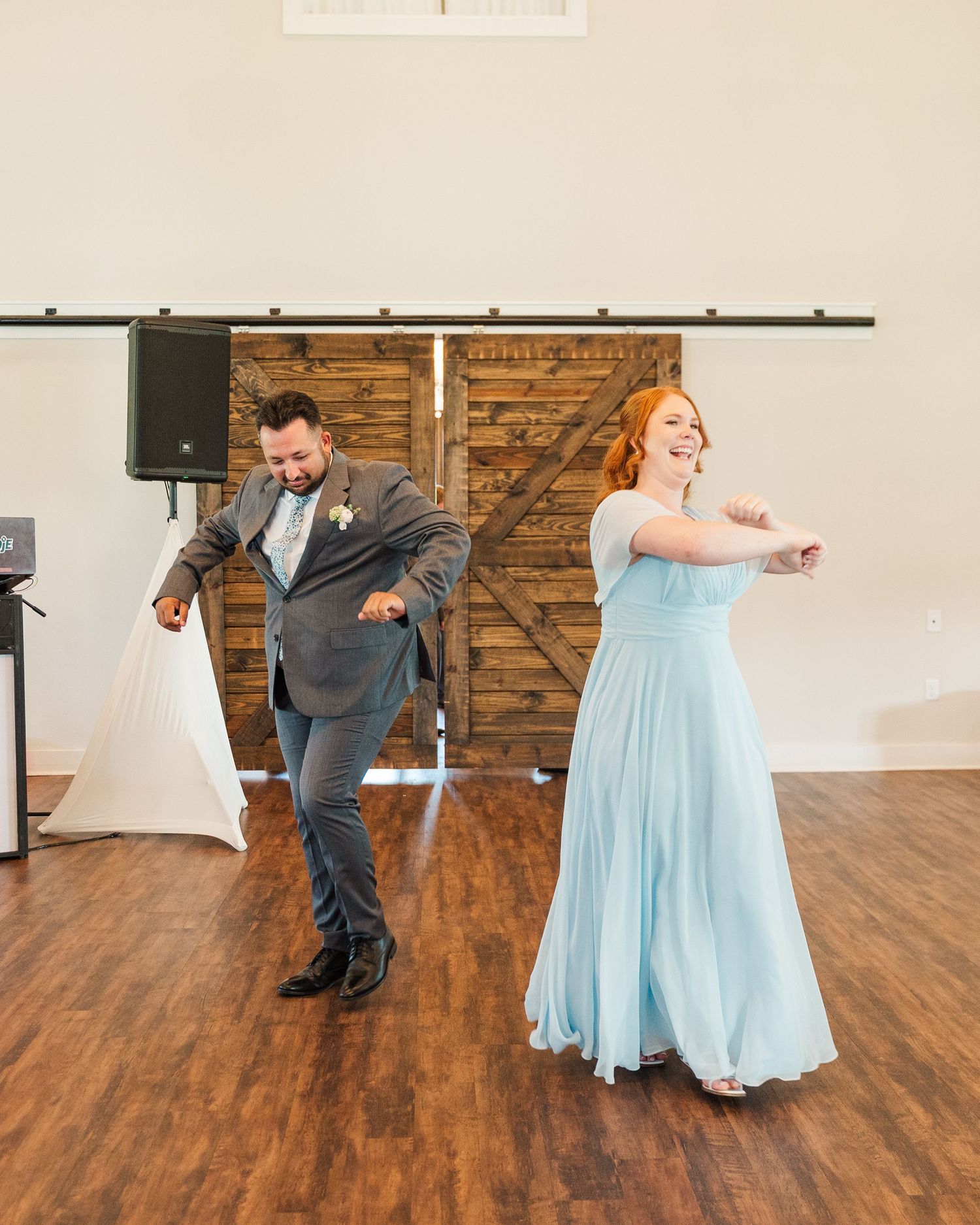 A man in a gray suit and woman in a light blue gown dance joyfully at a rustic barn wedding reception.