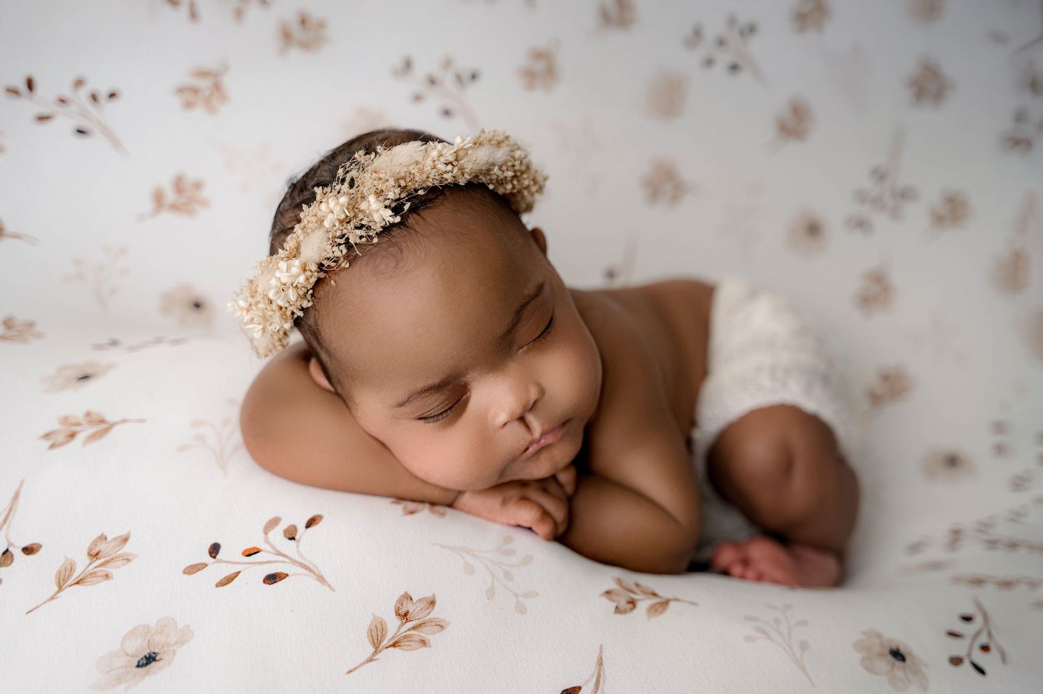 Newborn baby sleeping peacefully on soft floral bedding while wearing a delicate lace headpiece.