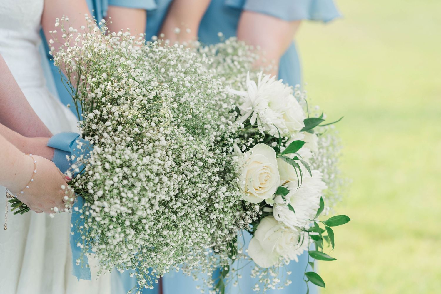 A row of bridesmaids in light blue dresses hold delicate bouquets of white and green flowers at an outdoor wedding.