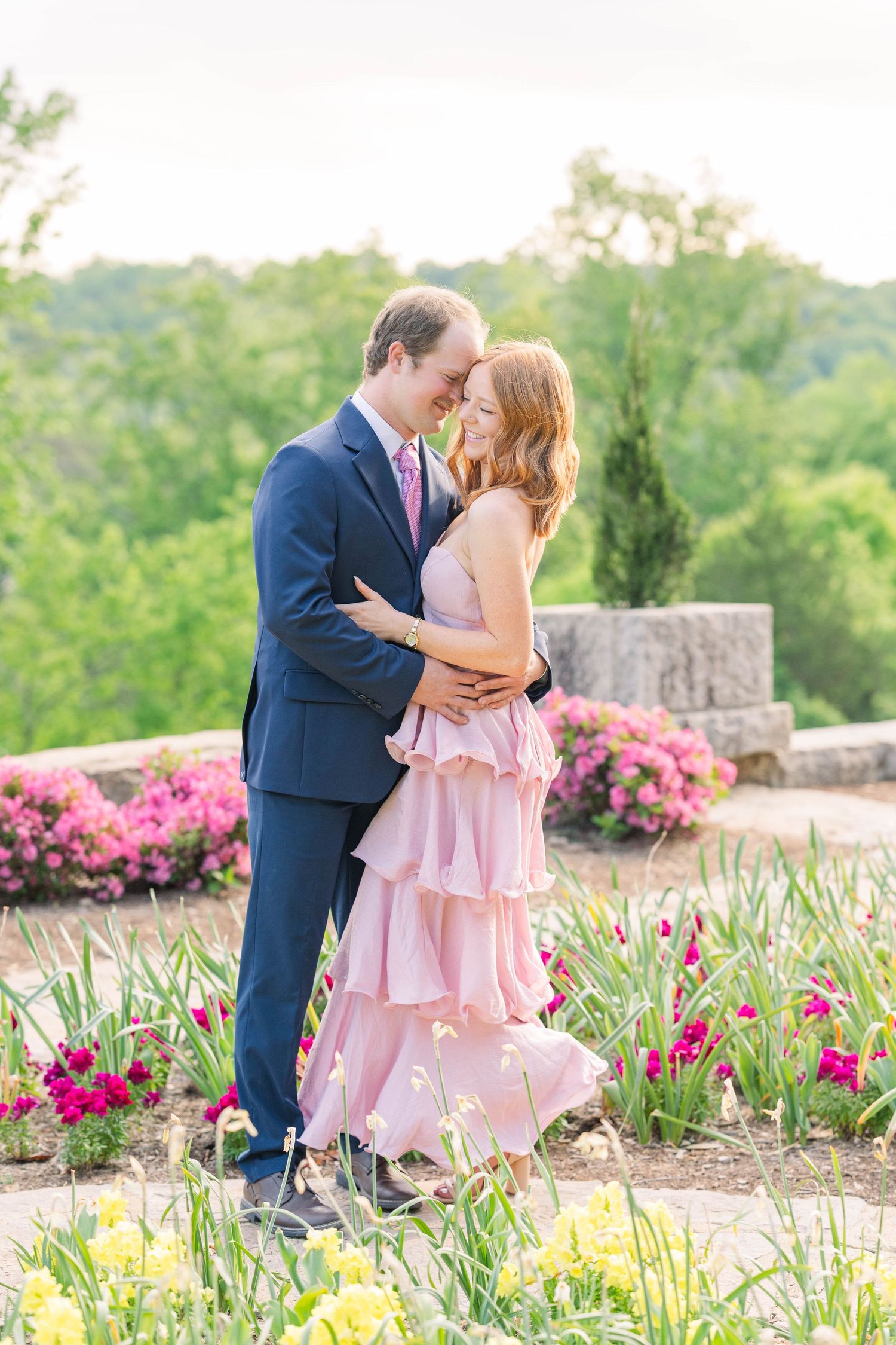 Couple embracing in a garden with pink tiered dress and navy suit, surrounded by colorful spring flowers.