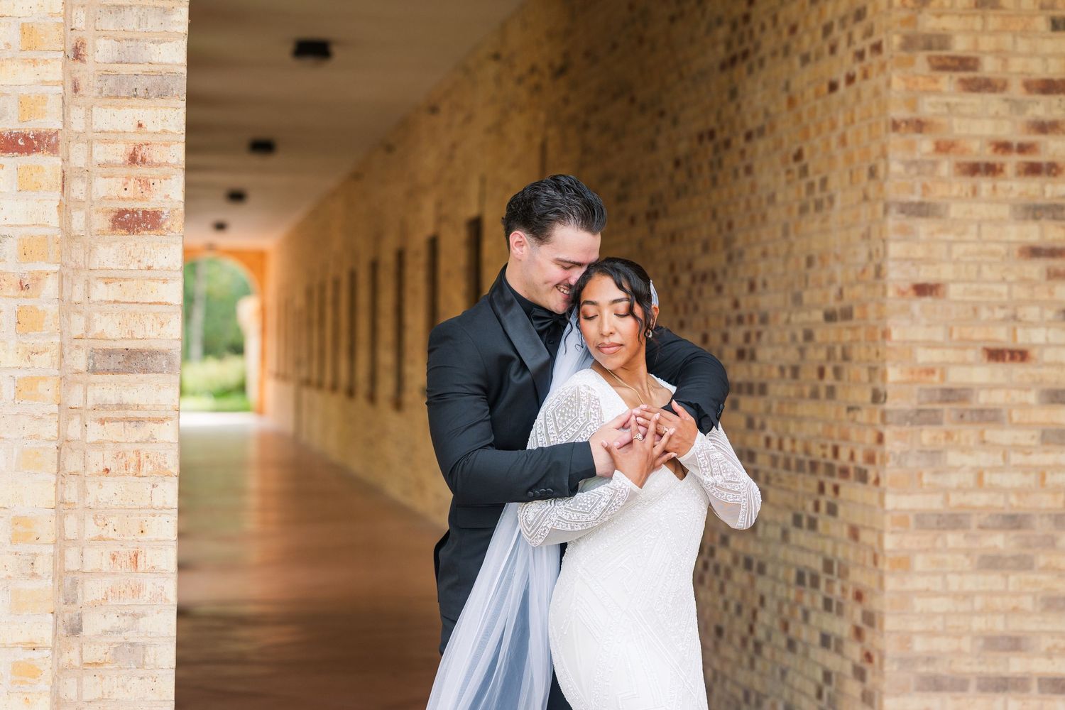 A groom in a black tuxedo embraces his bride in a white lace gown under a brick archway corridor.