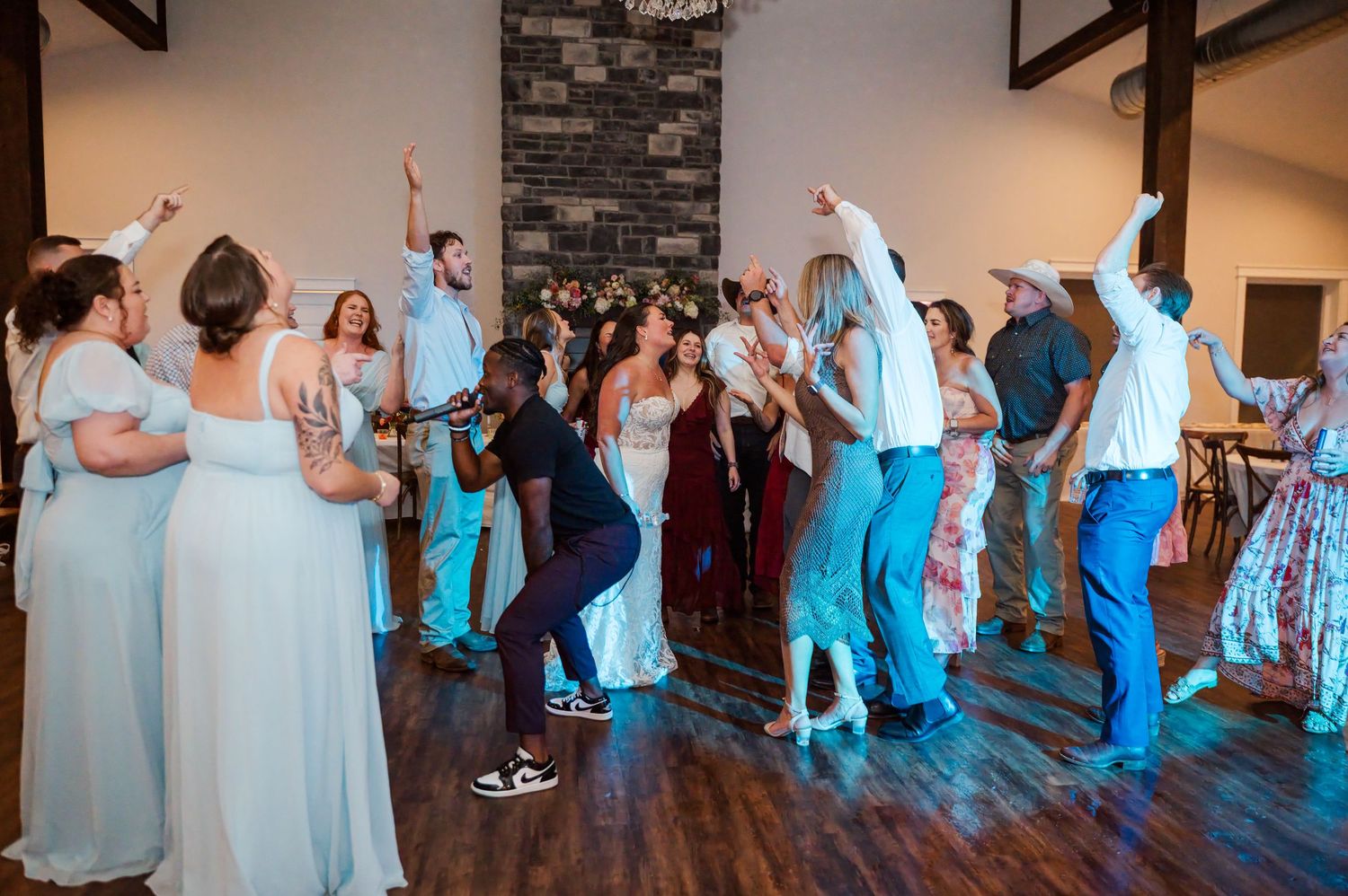 Wedding guests in formal attire dance and cheer with raised arms on a wooden dance floor in a rustic venue with a stone fireplace.