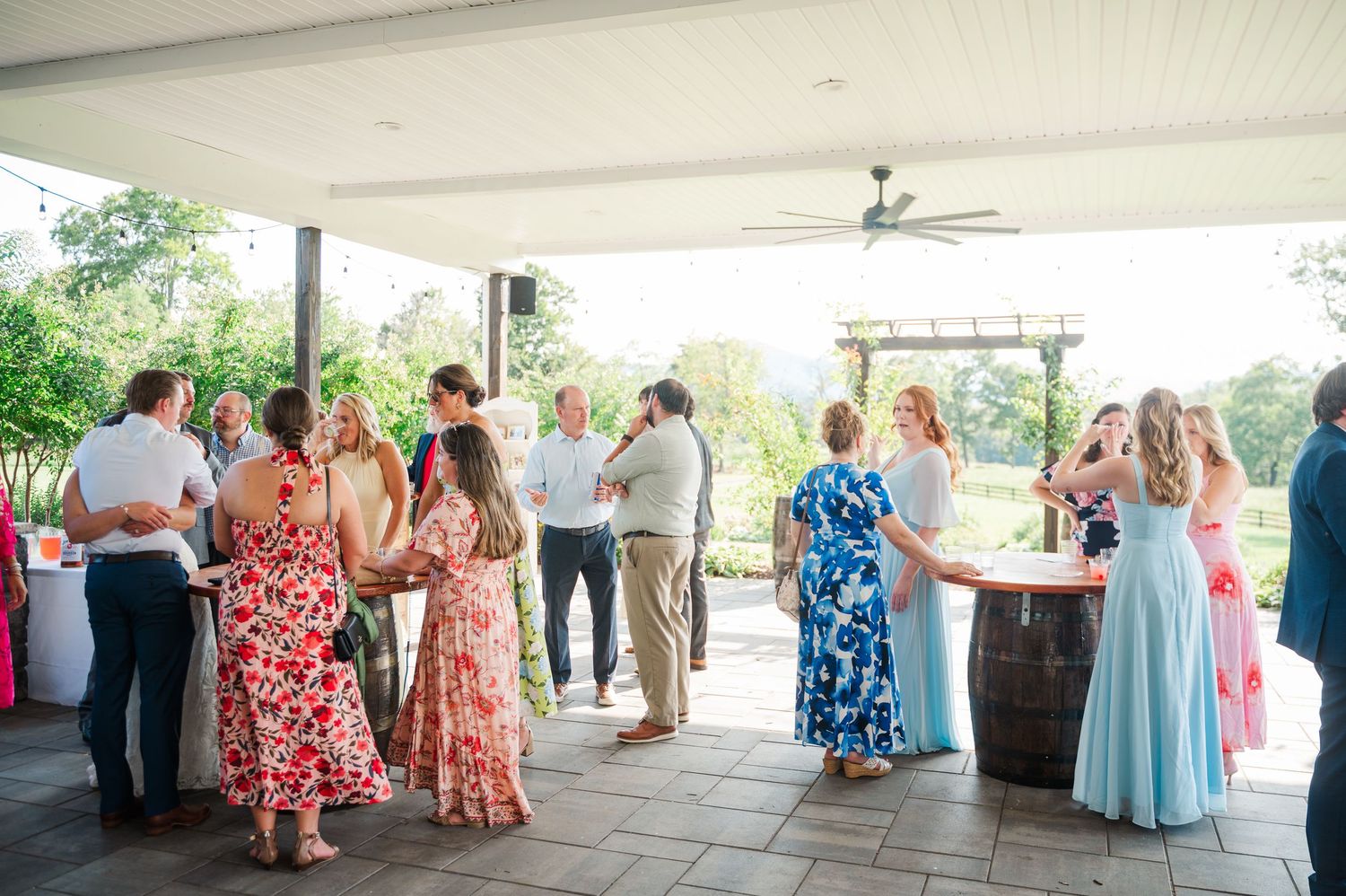 Wedding guests mingle on a covered outdoor patio with barrel tables during a sunny summer reception.
