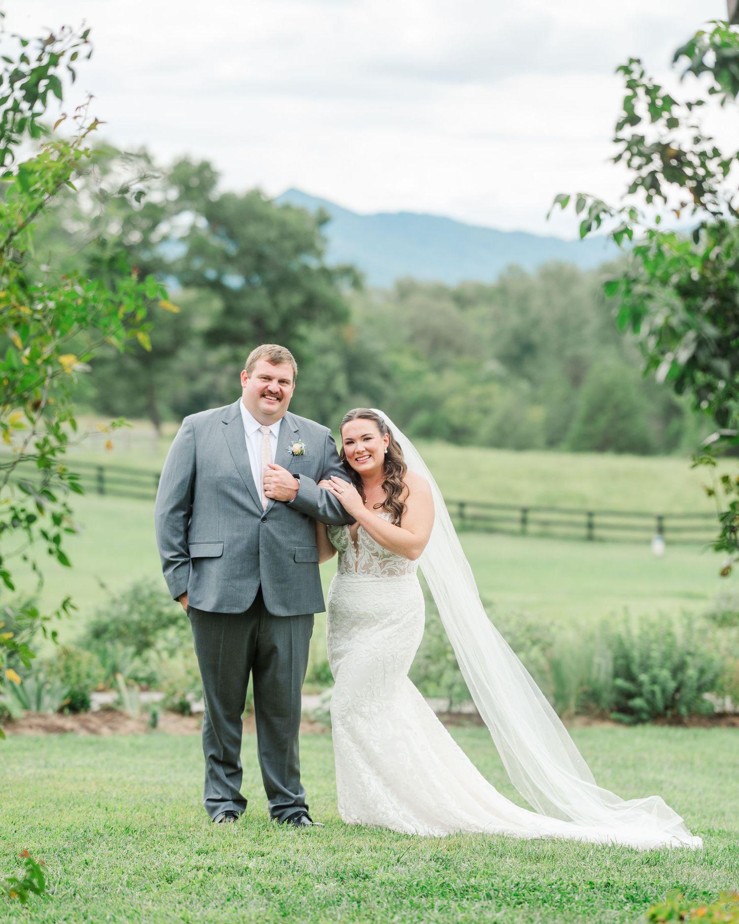 Bride in lace gown and veil stands with groom in gray suit on green lawn with mountain backdrop.
