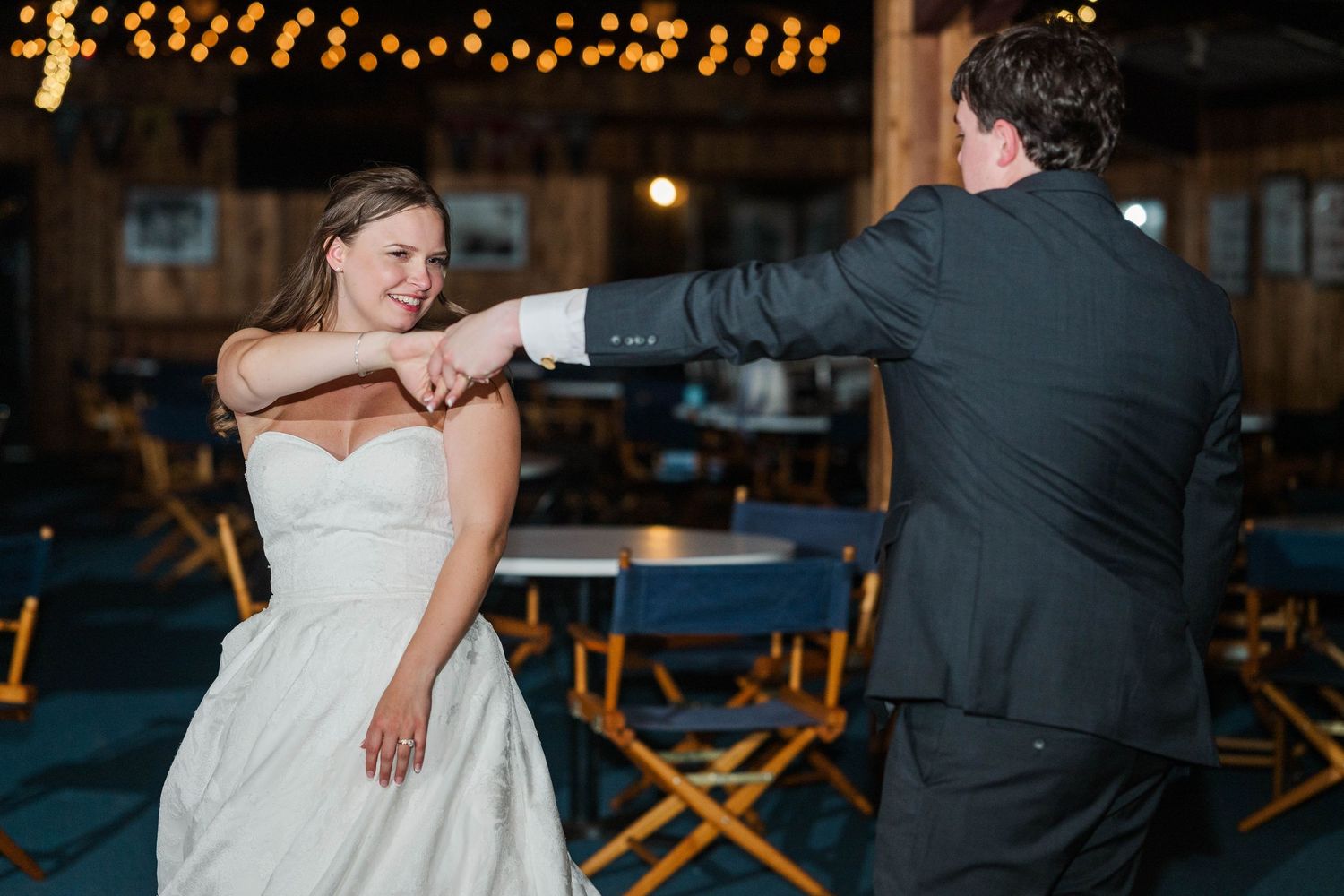 Couple dancing at wedding reception with string lights glowing in rustic wooden venue.