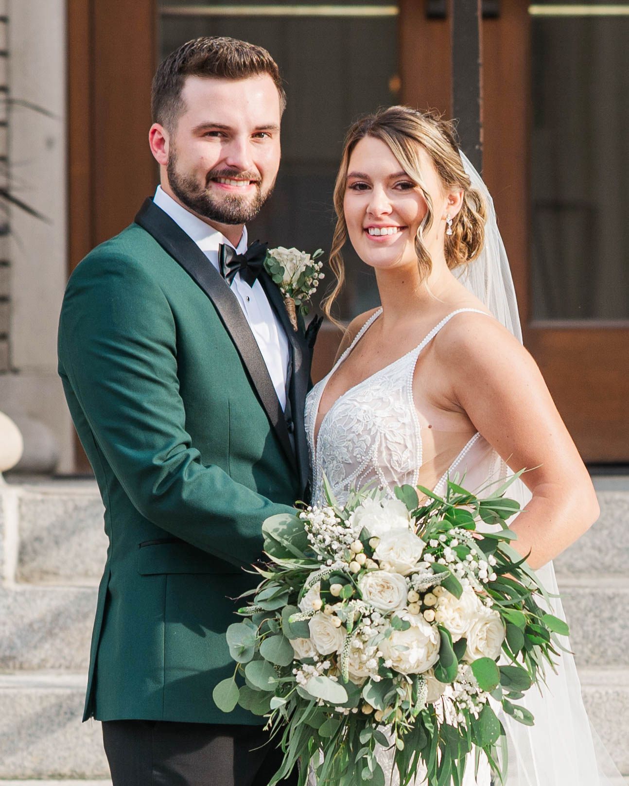 Wedding couple poses together with elegant white and green floral bouquet against stone backdrop.