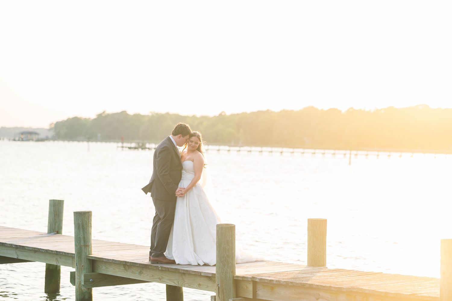 A romantic sunset photoshoot of a couple in wedding attire on a wooden dock by the water.