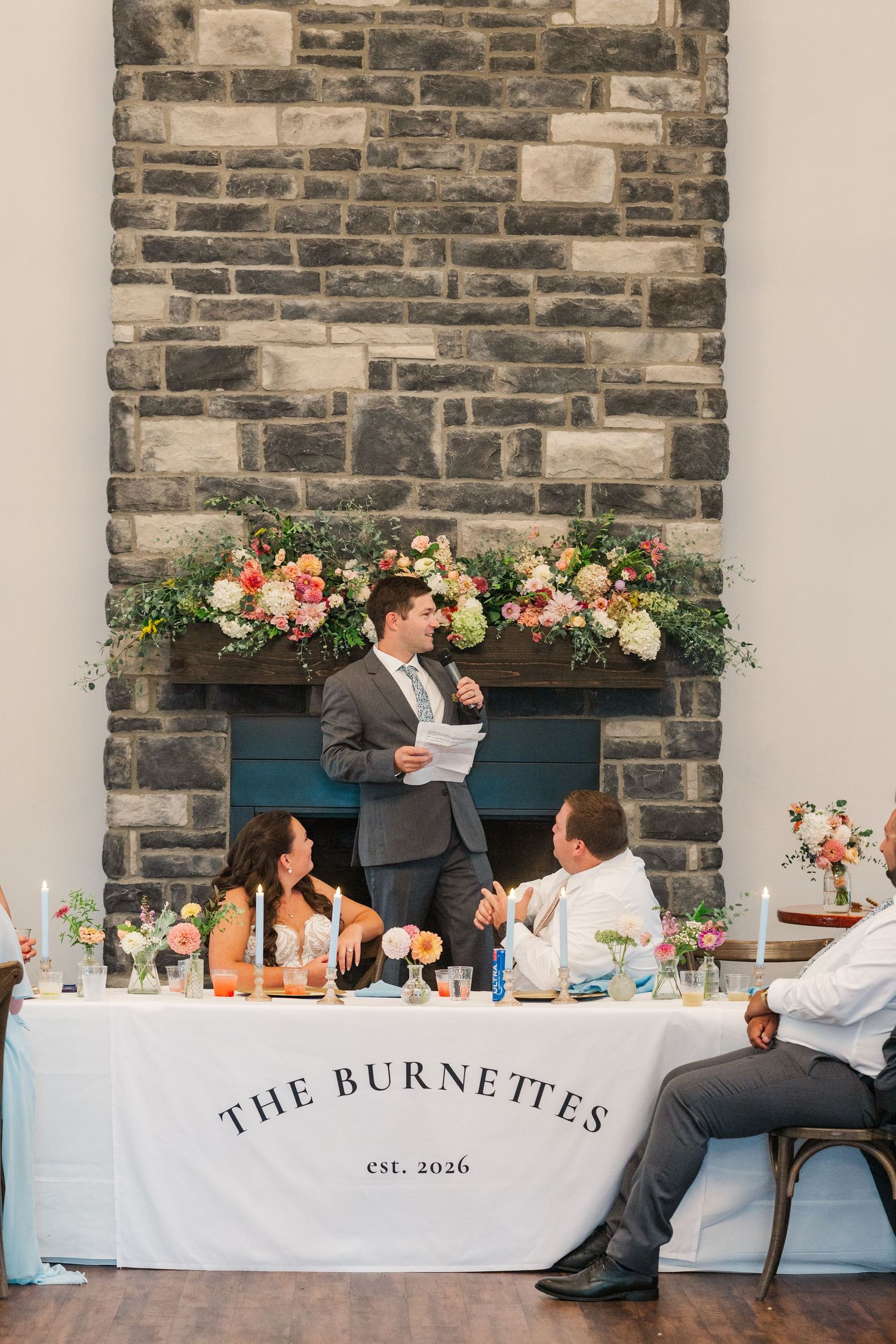 Best man gives toast at The Burnettes wedding sweetheart table in front of a stone fireplace.