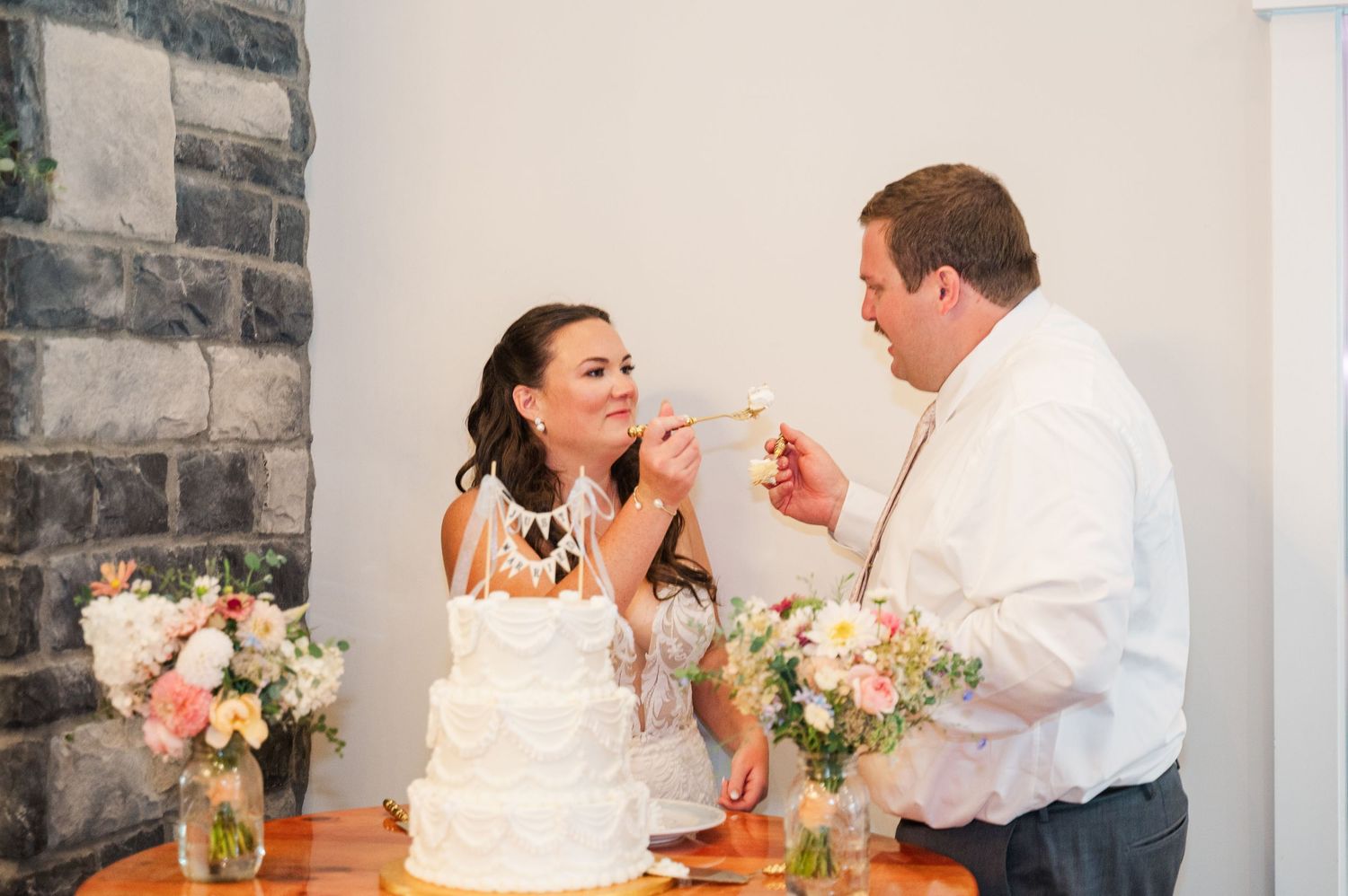 Bride and groom share sweet cake cutting moment at elegant wedding reception with floral arrangements nearby.