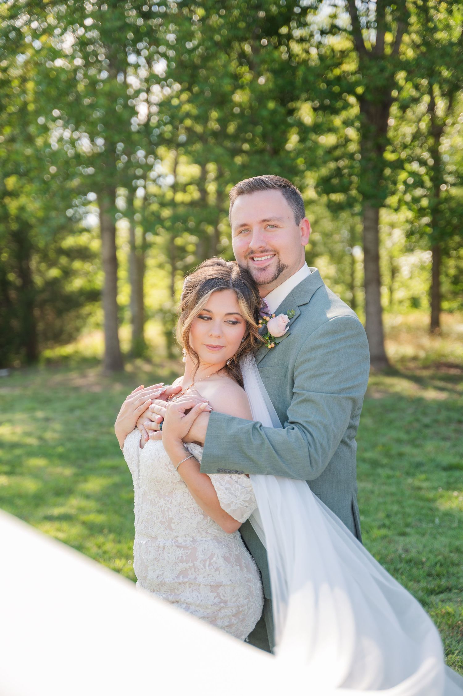 Bride and groom embrace outdoors in lush green setting, she in lace gown with flowing veil, he in sage suit with boutonniere.