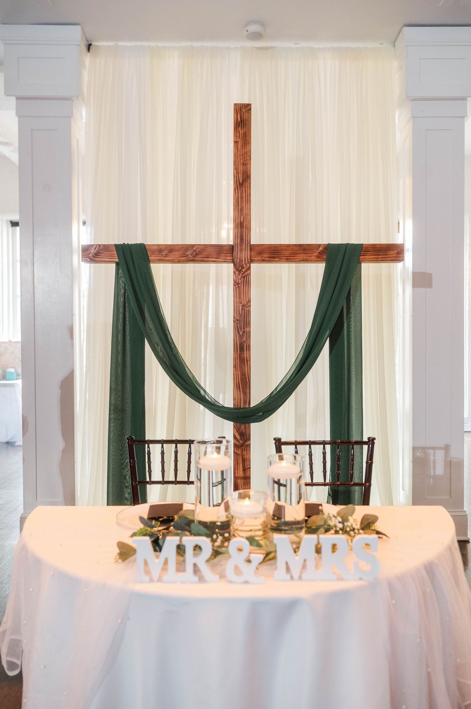 Wedding reception head table with white linens and emerald green draping behind wooden cross.