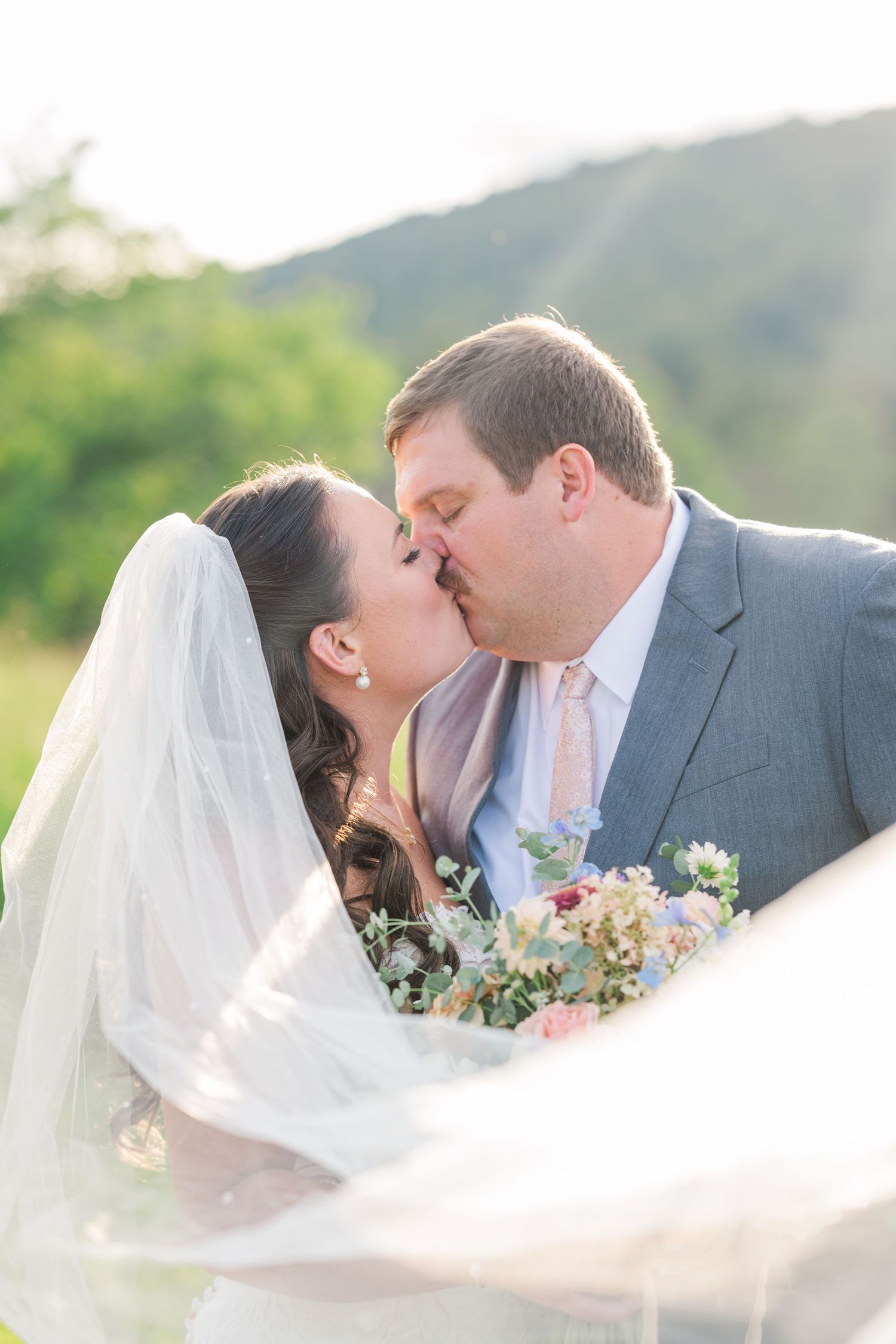Bride and groom kissing outdoors with a flowing veil and lush green hills in the background.