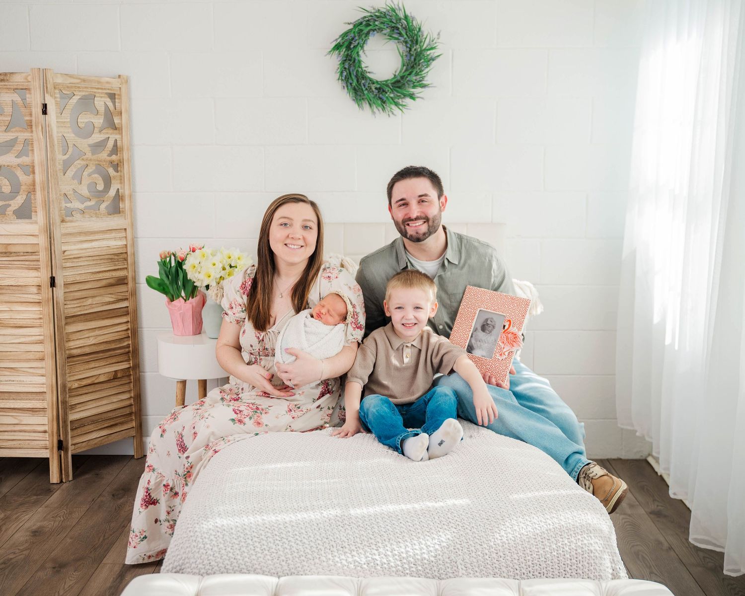 Happy family of four poses on a white bed with newborn baby in a bright farmhouse-style room with wreath decor.
