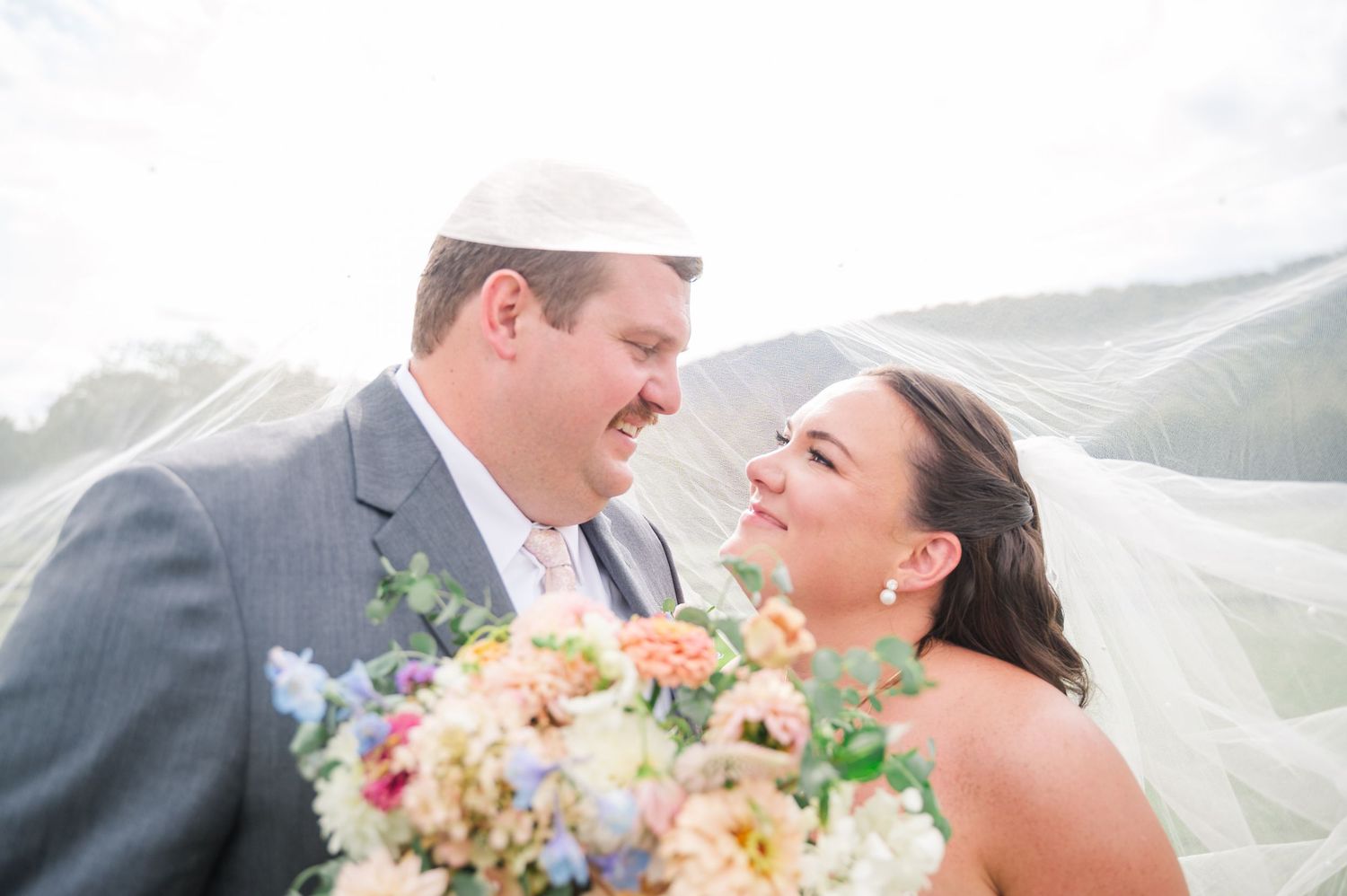 Happy couple sharing a romantic moment under a flowing bridal veil with a colorful wedding bouquet.