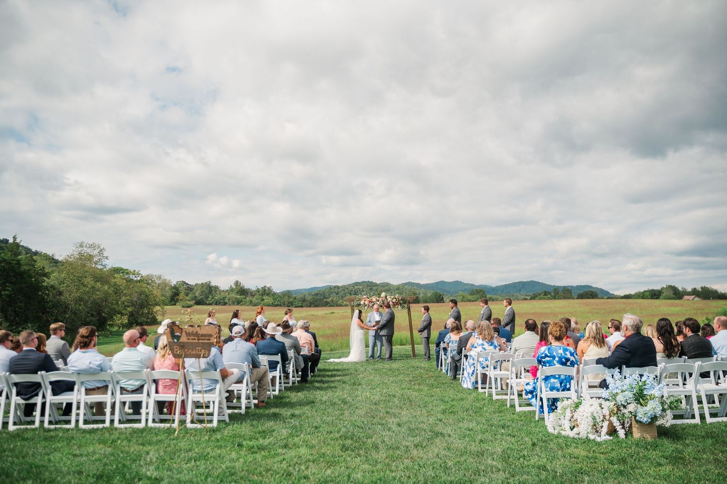 Outdoor wedding ceremony with guests seated in white chairs on a green lawn with mountain views.