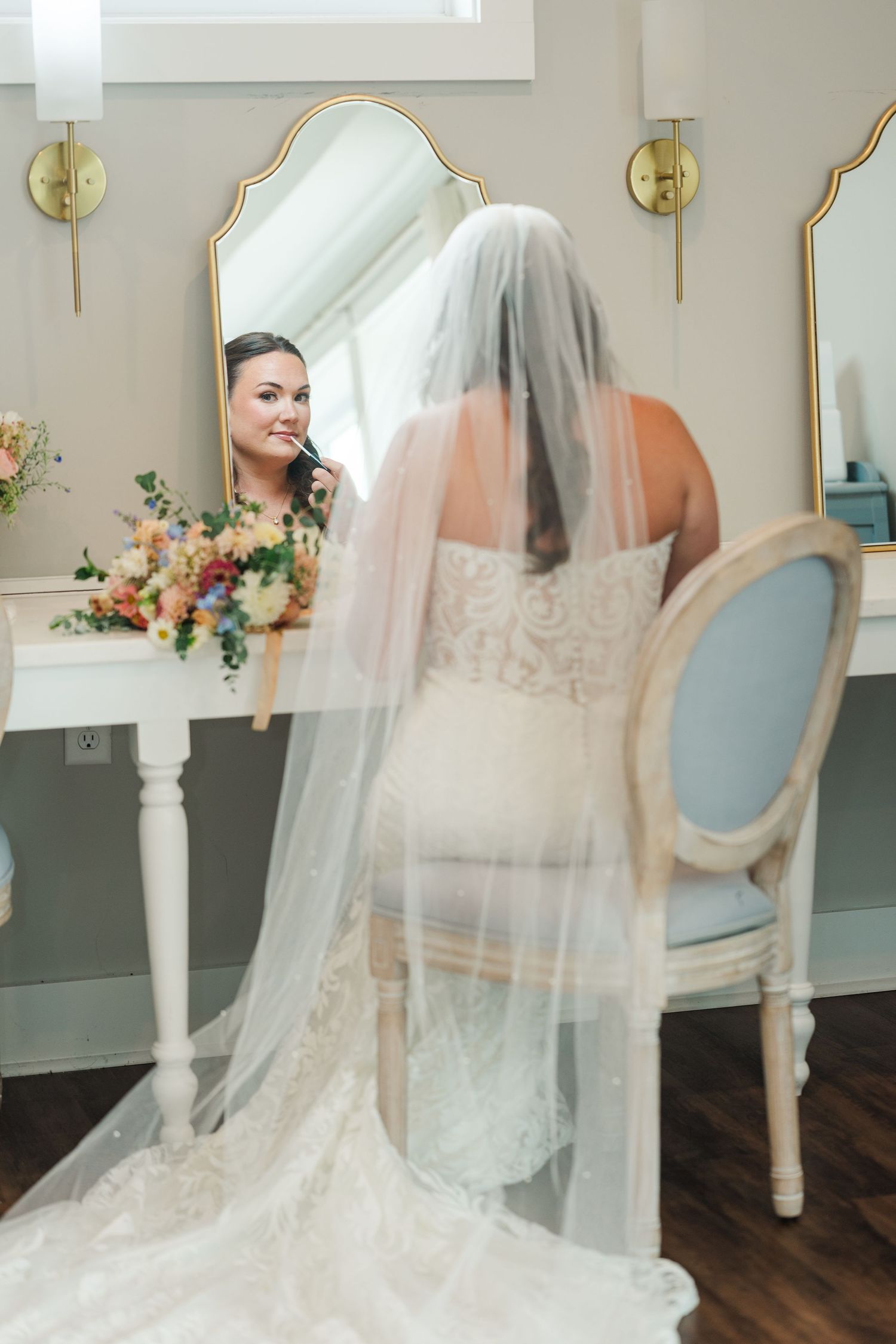 Bride in lace gown and veil sits at vanity mirror applying makeup before her wedding ceremony.