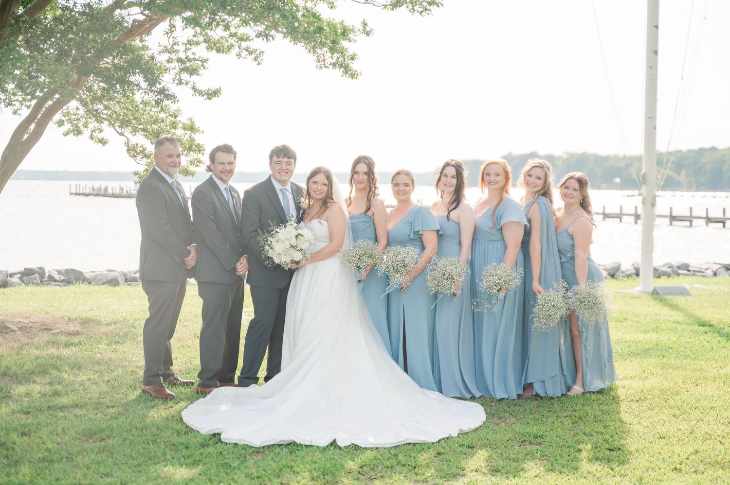 Wedding party in light blue dresses and dark suits poses together under a large tree by the water.