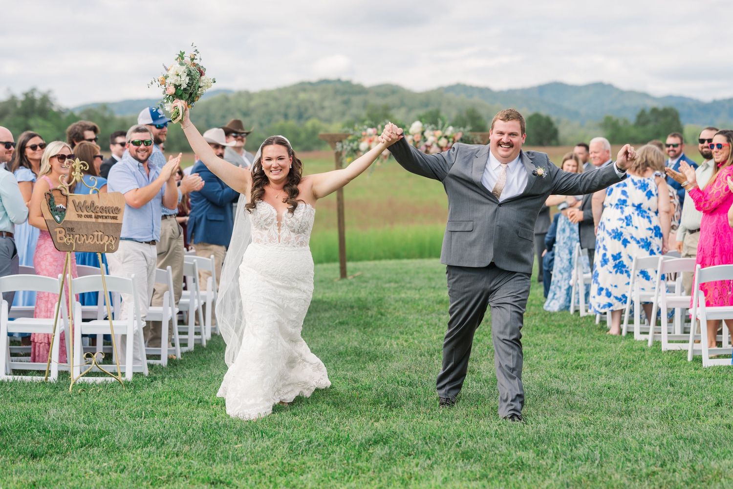Joyful newlyweds walk down outdoor wedding aisle, arms raised, guests cheering, mountains and green fields in background.