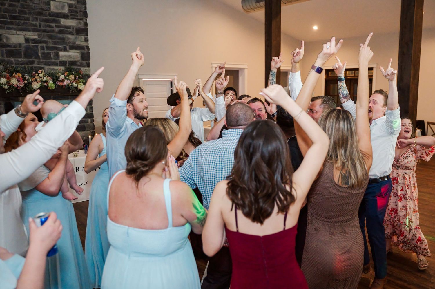 Wedding guests in formal attire dance with raised arms and cheer together on a crowded dance floor at a lively reception.