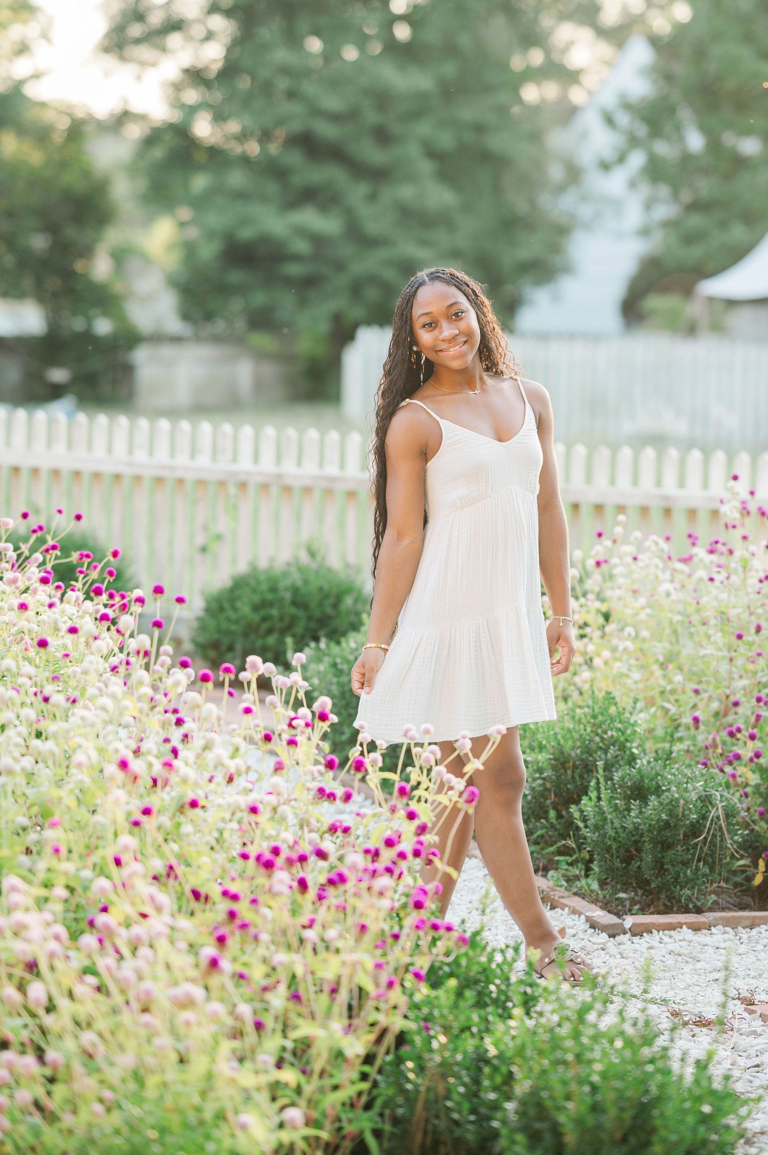 Young woman in white sundress smiling among pink wildflowers in a garden with white picket fence.