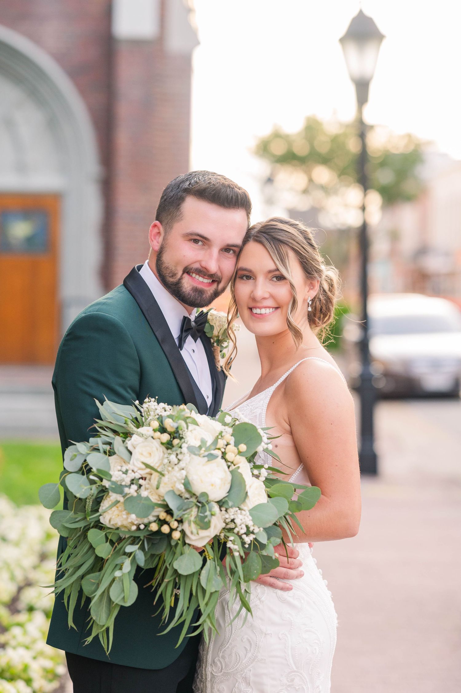 A couple poses together outdoors with a lush white and green floral bouquet.