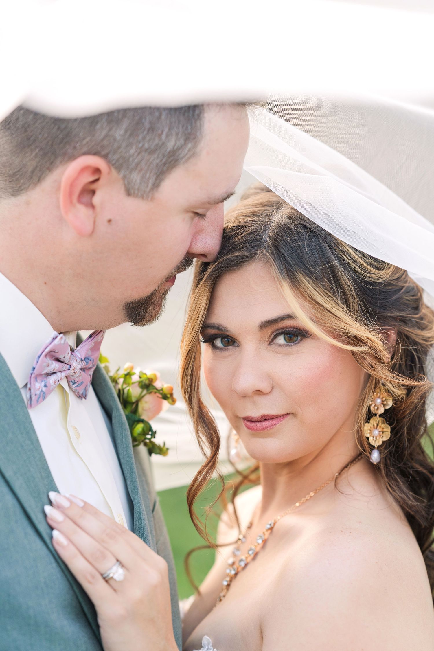 Bride and groom share an intimate moment under a veil, bride wearing gold earrings and necklace on wedding day. Showcasing the wedding band.