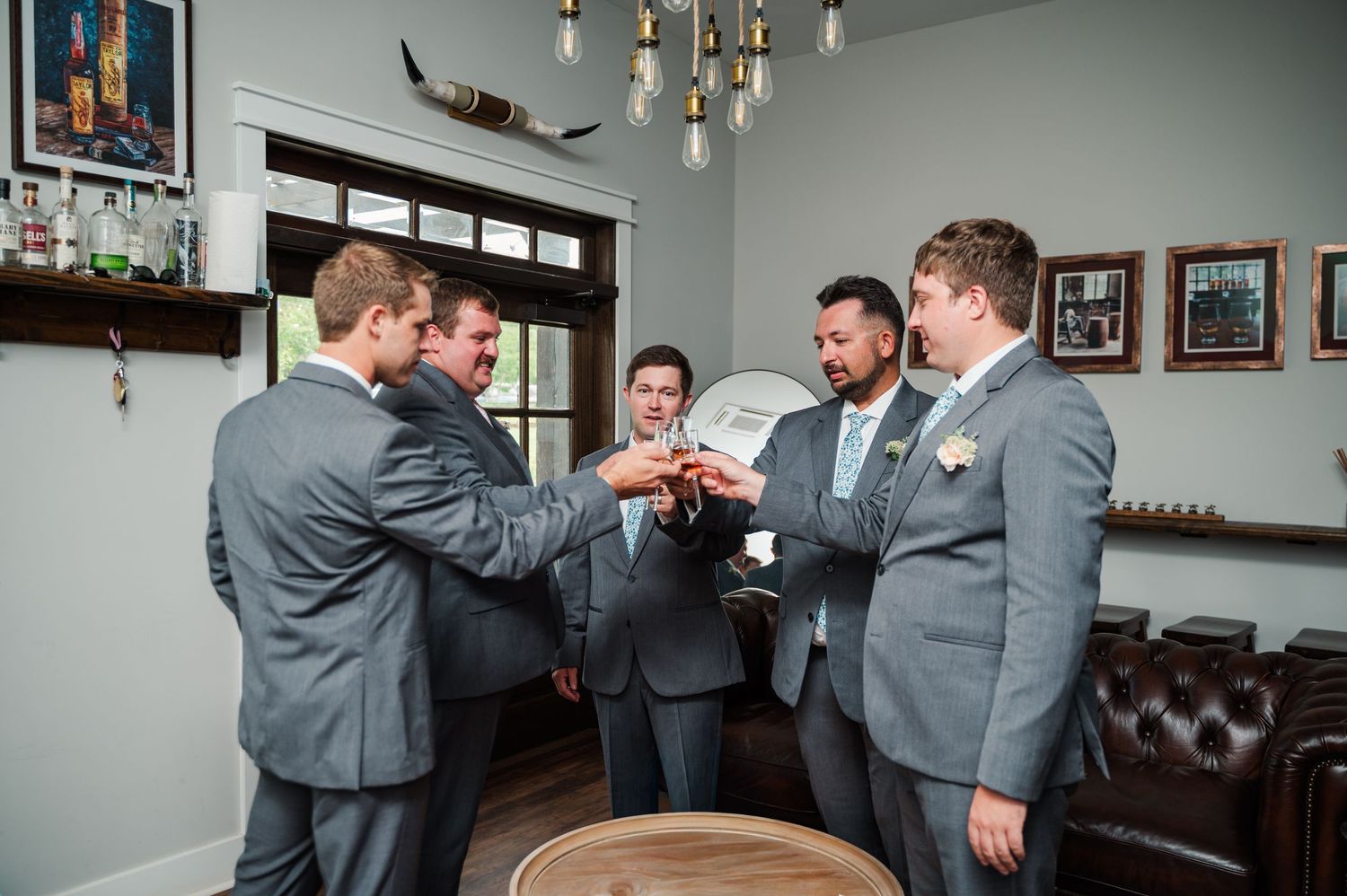 Groomsmen in gray suits gather around groom holding wedding cake topper in elegant venue before ceremony.