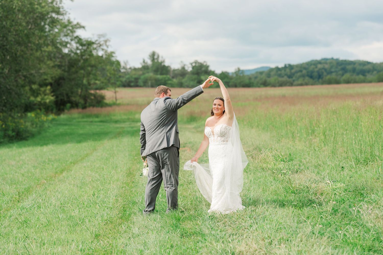 Groom twirls bride in white wedding gown and veil in a lush green field with mountains behind.