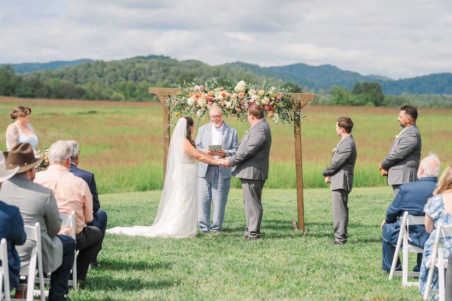 Bride and groom exchange vows under a wooden arch adorned with peach and pink flowers at an outdoor wedding.