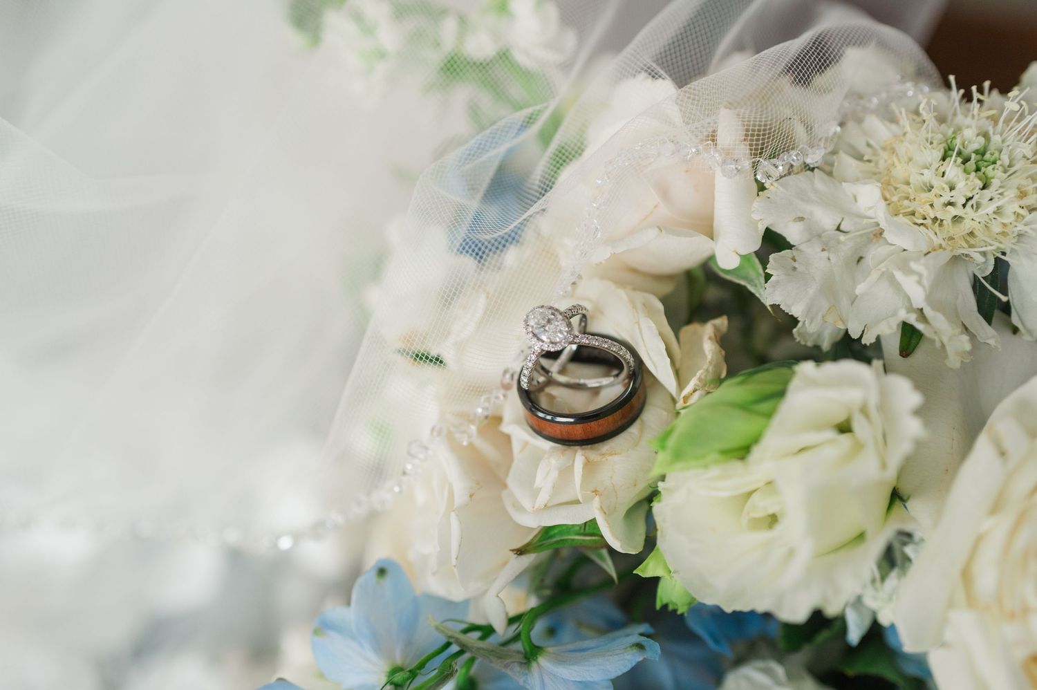 Wedding rings resting on a bridal bouquet of white and blue flowers with a delicate veil backdrop.