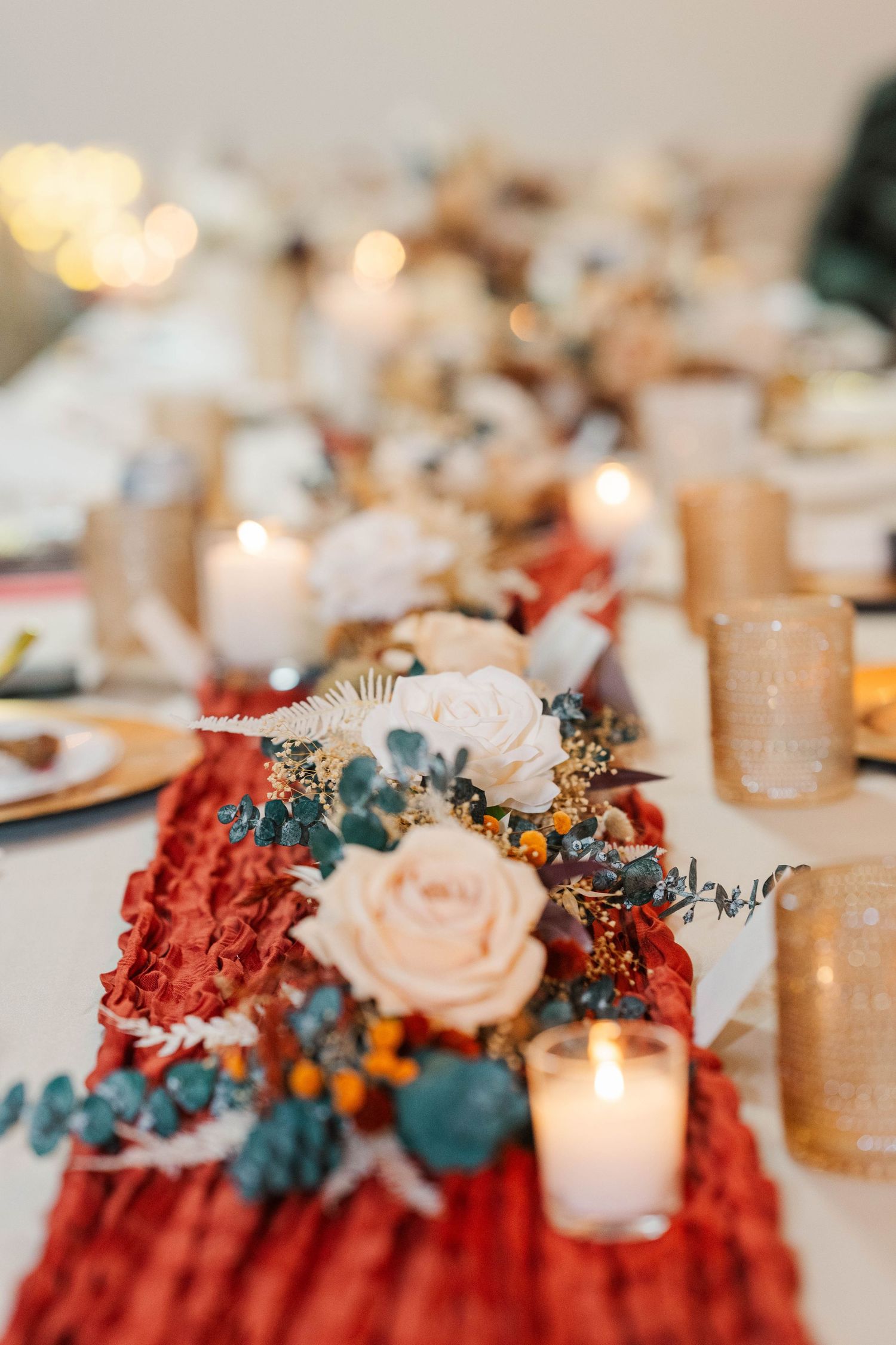 Elegant wedding table centerpiece featuring blush roses, burgundy flowers, and teal accents with glowing candles.
