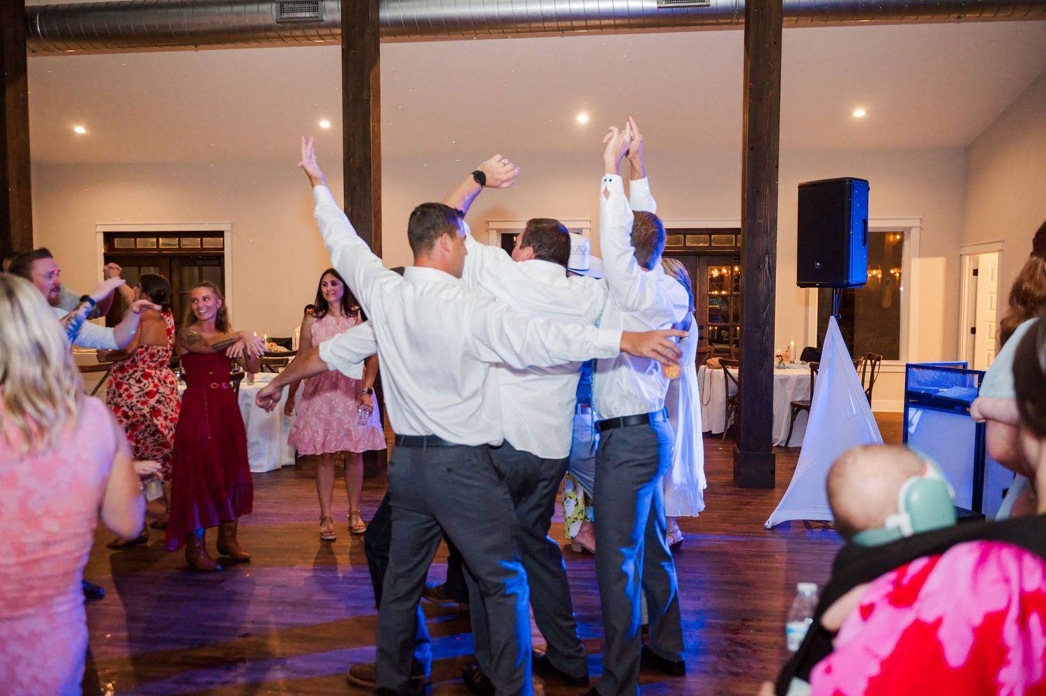 Wedding guests in white shirts dance with raised arms on a wooden dance floor in a rustic reception hall while others watch.