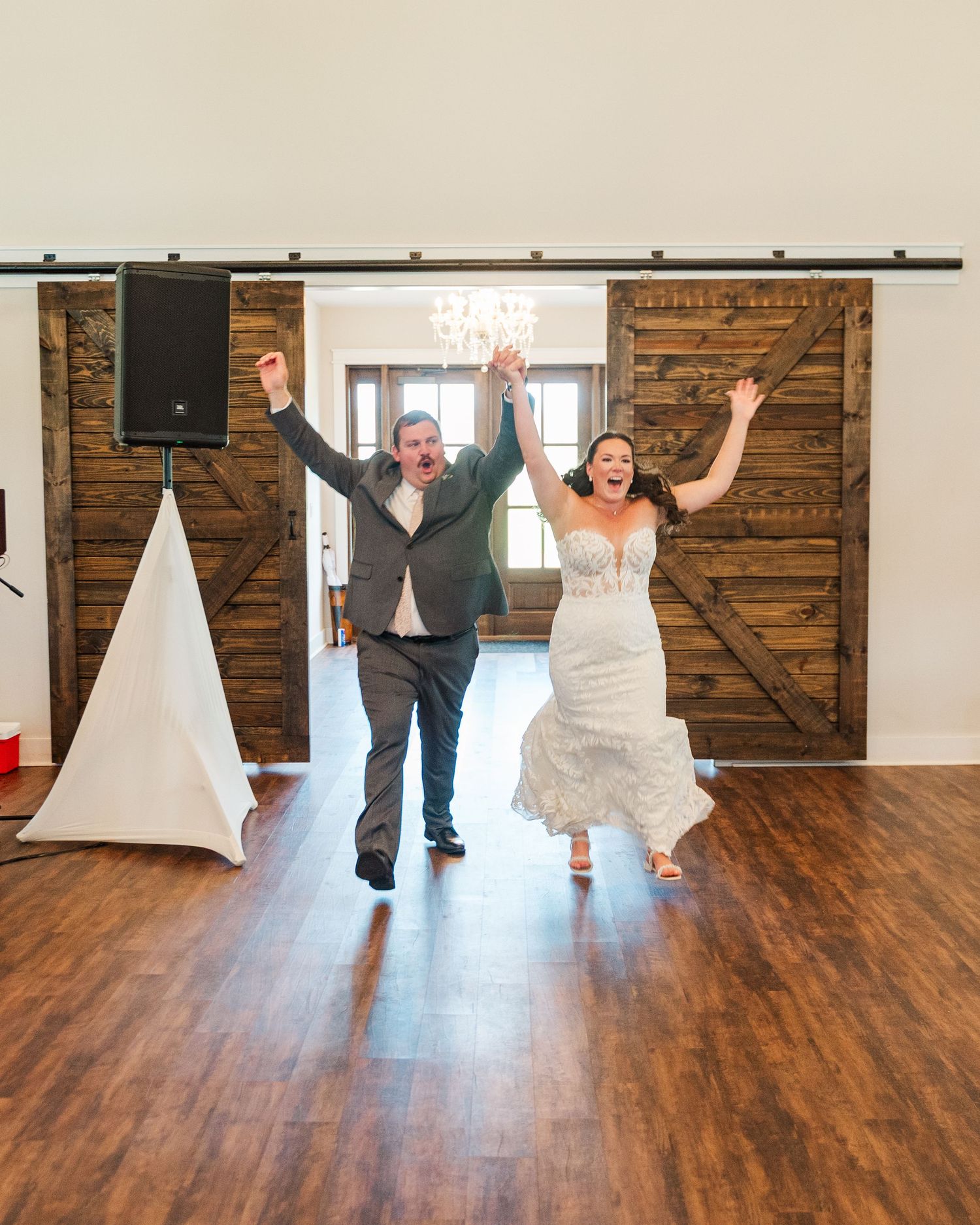Joyful newlyweds make grand entrance at rustic barn wedding reception, raising hands in celebration on hardwood dance floor.