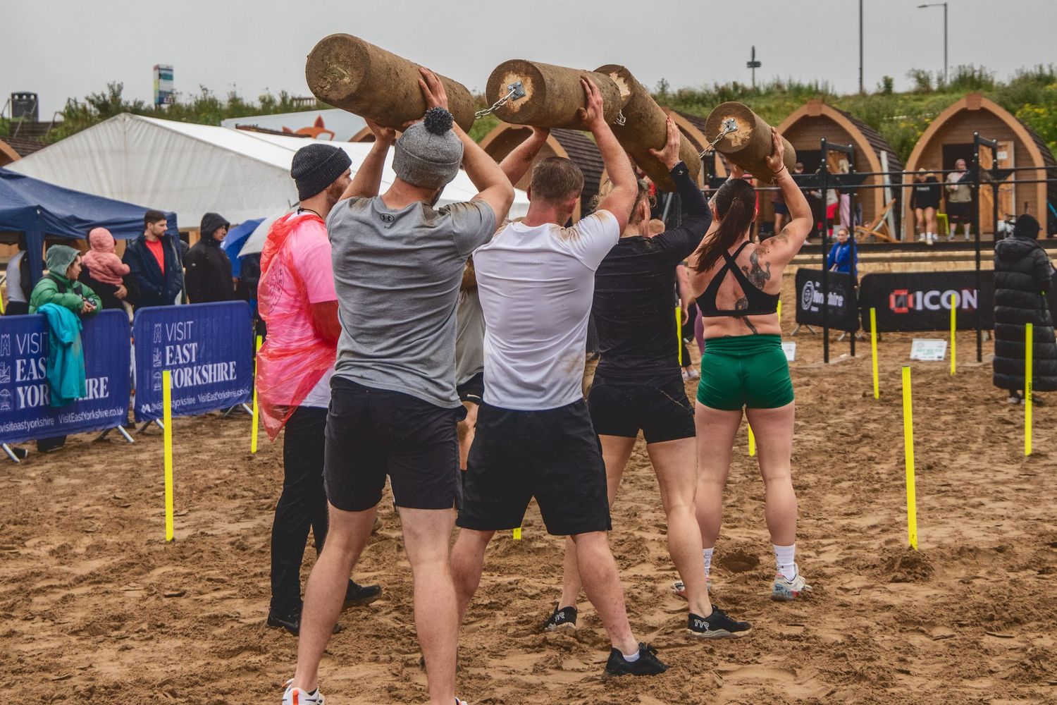 Athletes compete in a log lifting event at an outdoor strongman competition on sandy ground with spectators watching.
