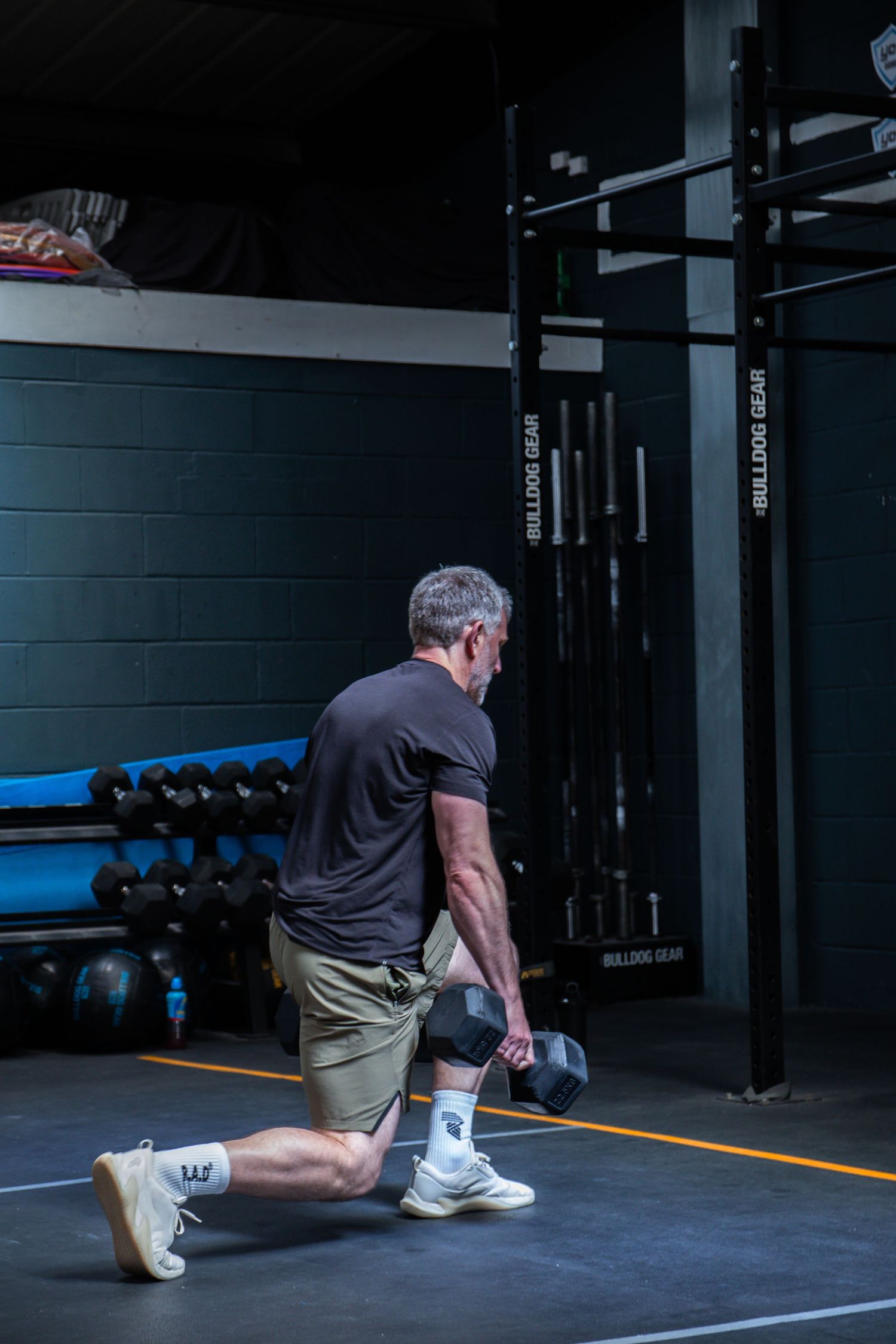 Athlete performs dumbbell lunges on black gym floor with weightlifting equipment and racks visible in background.